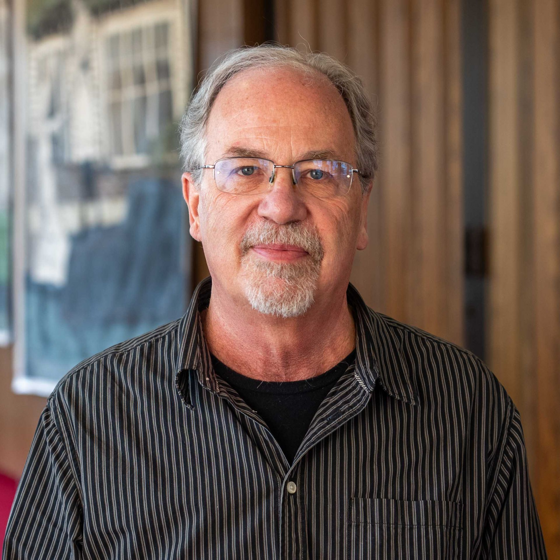 Portrait of a man with glasses and gray hair, wearing a striped shirt, standing indoors.