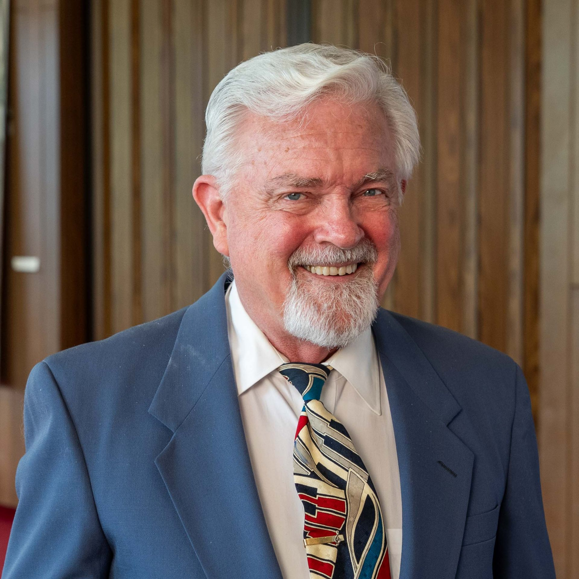 Smiling man in a blue suit and patterned tie standing before wood paneling