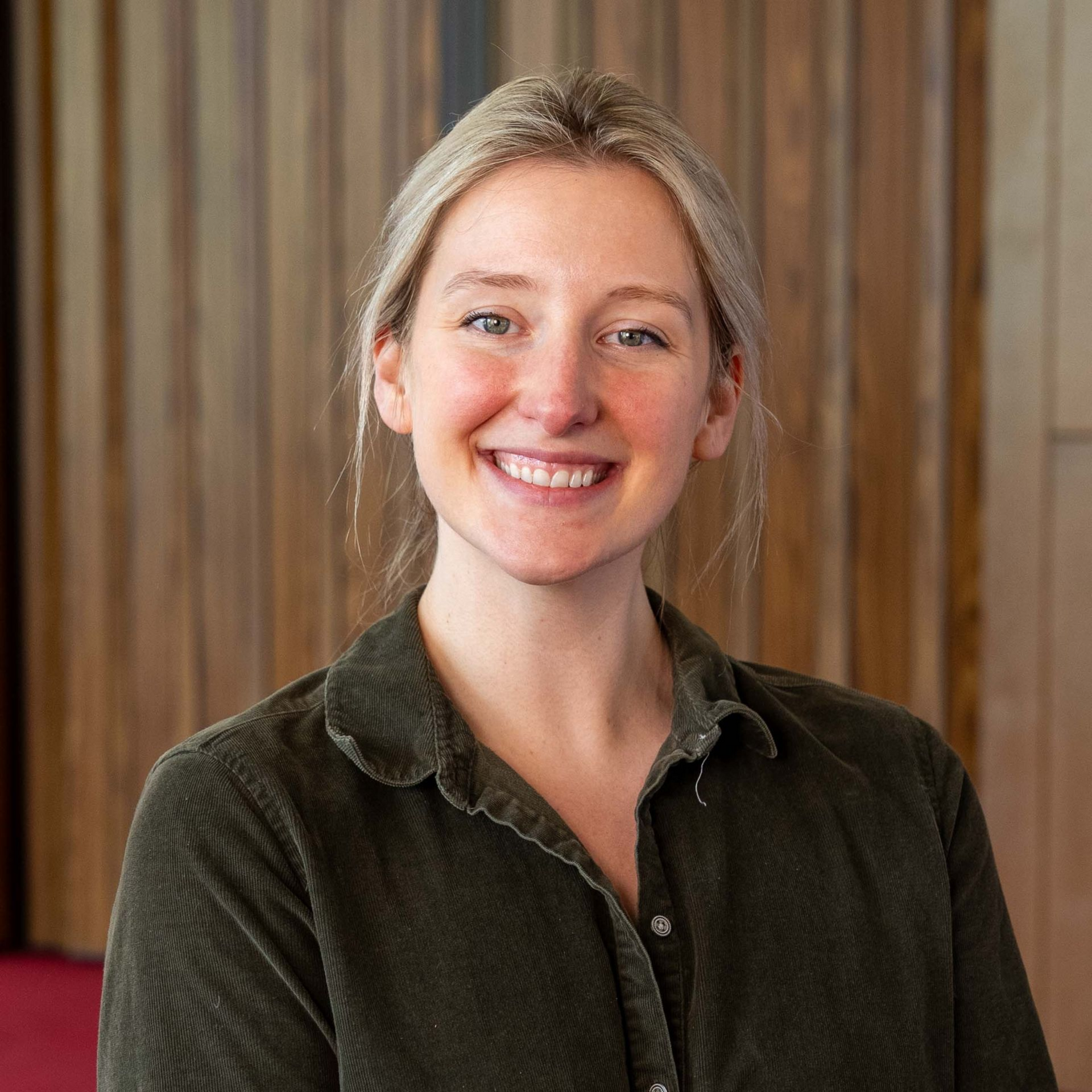 Smiling person in a dark green shirt standing before a wood-paneled background