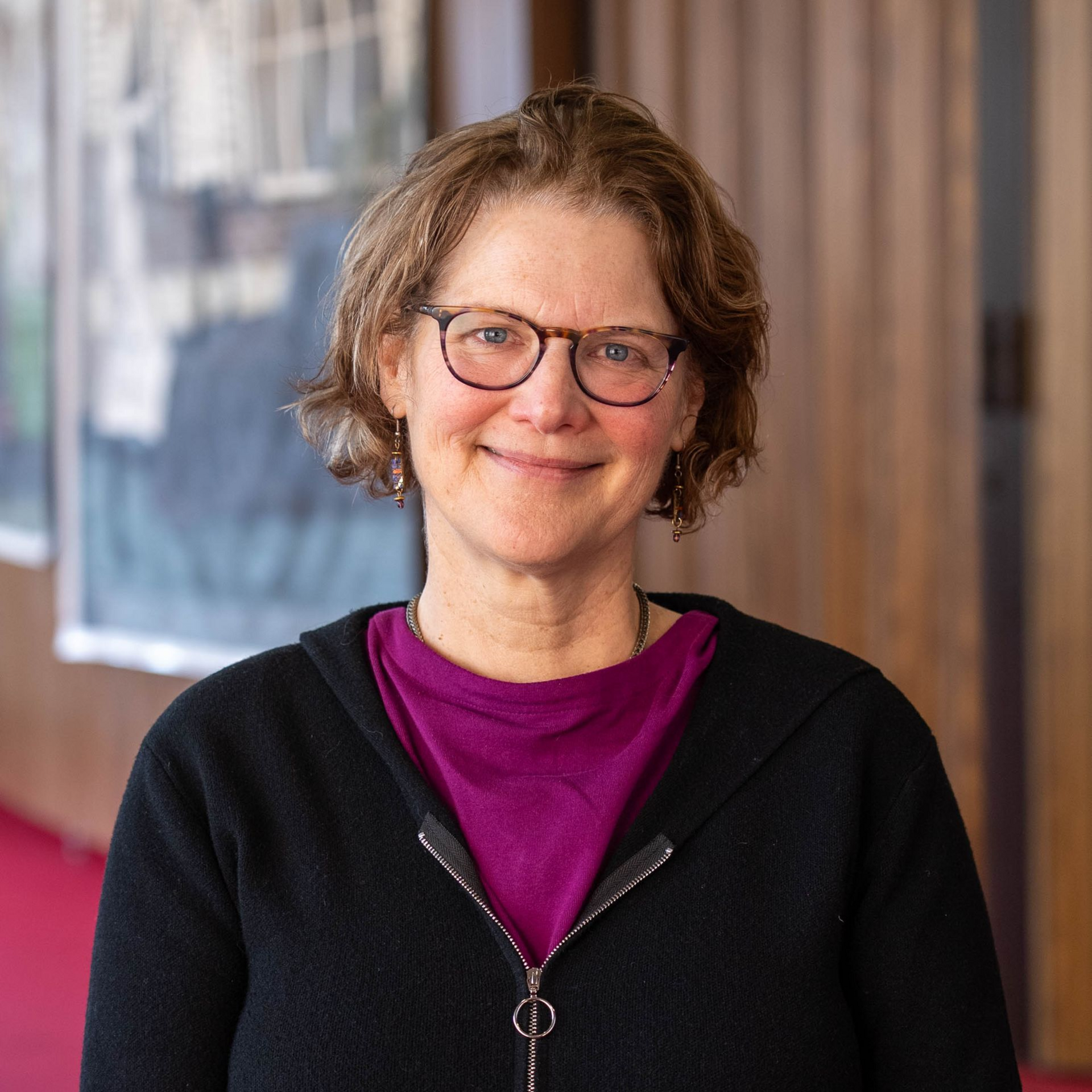 Smiling person in glasses and black sweater standing in a hallway with blurred wall art and wood paneling