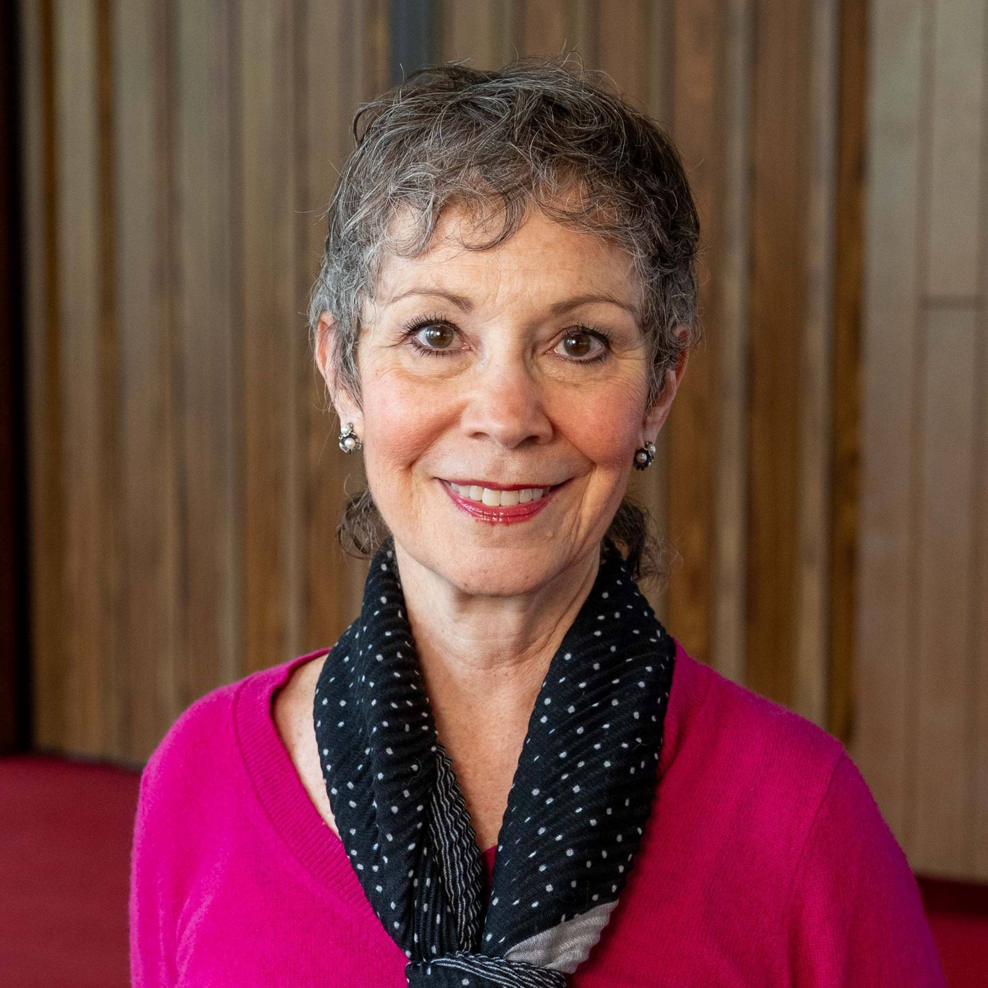 Smiling woman in a bright pink top and black polka-dot scarf against a wood-paneled background