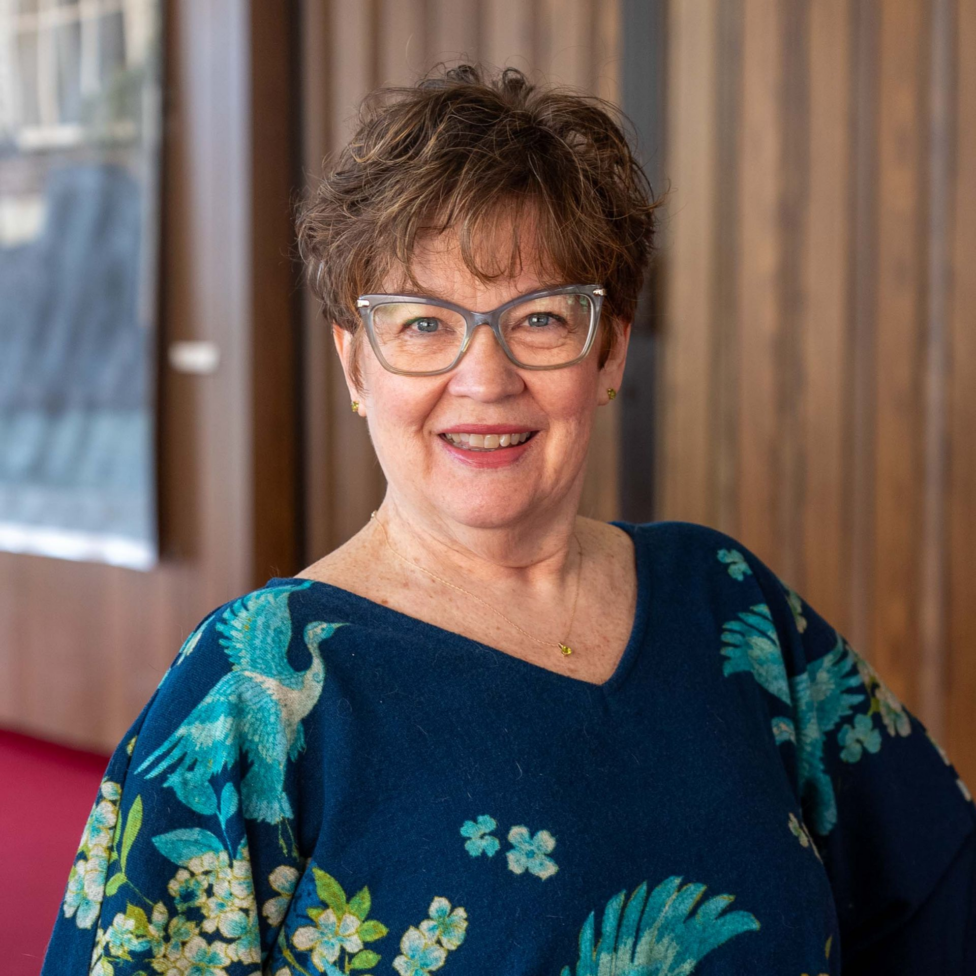 Smiling woman in glasses and a blue floral top, standing indoors against a wood-paneled wall