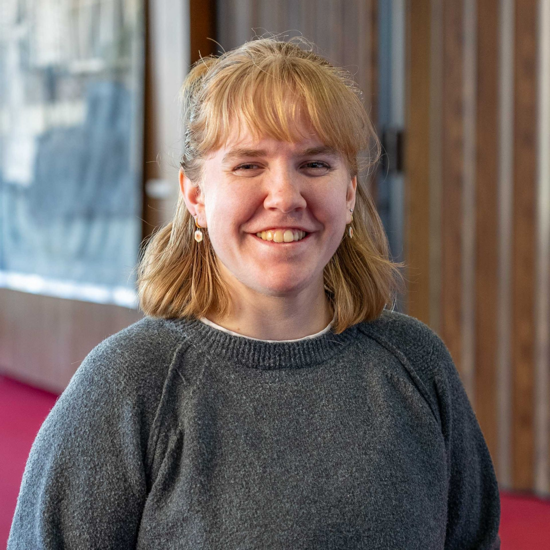 Smiling person in a dark gray sweater standing indoors by a wooden wall.