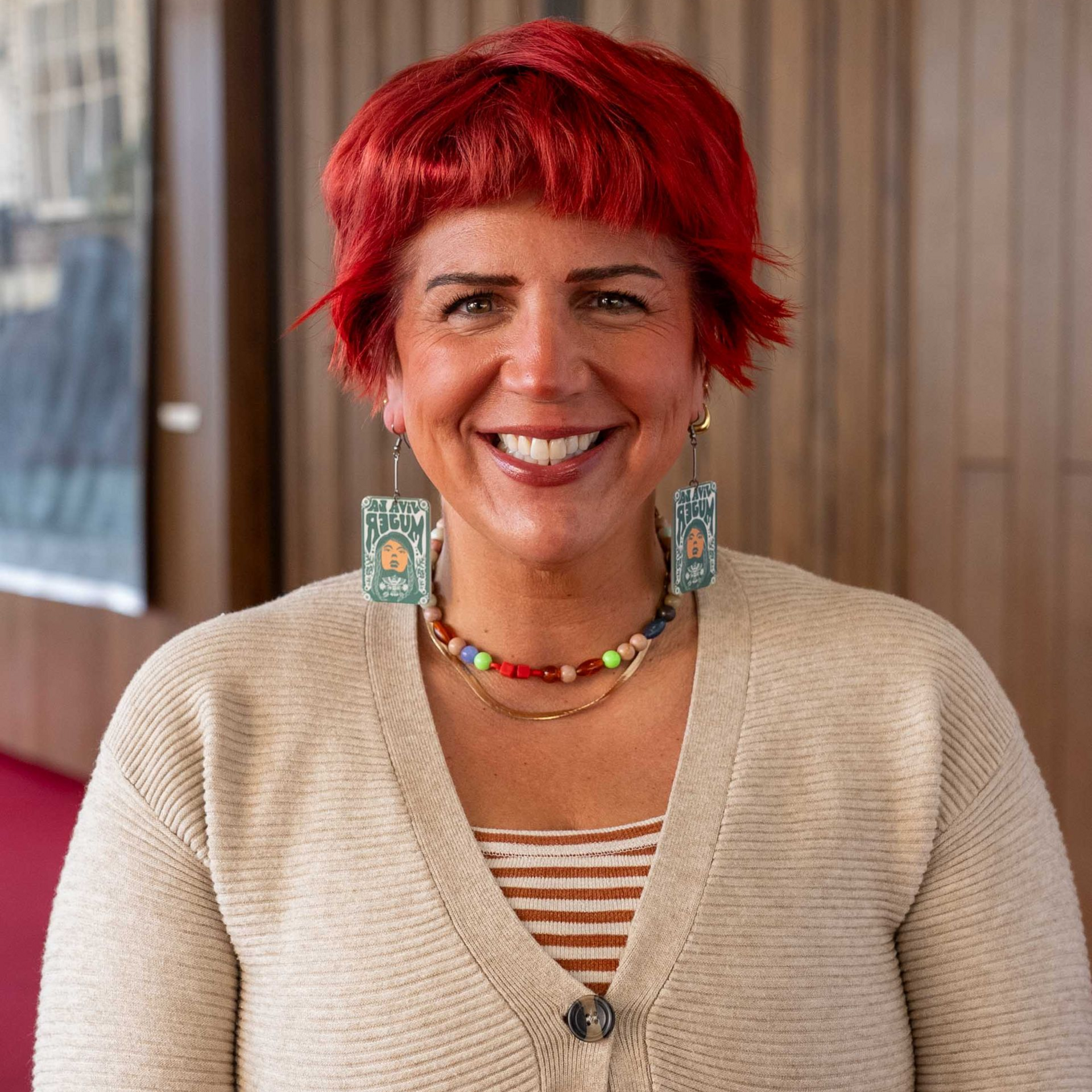 Smiling person with bright red hair, turquoise earrings, and a cream cardigan indoors