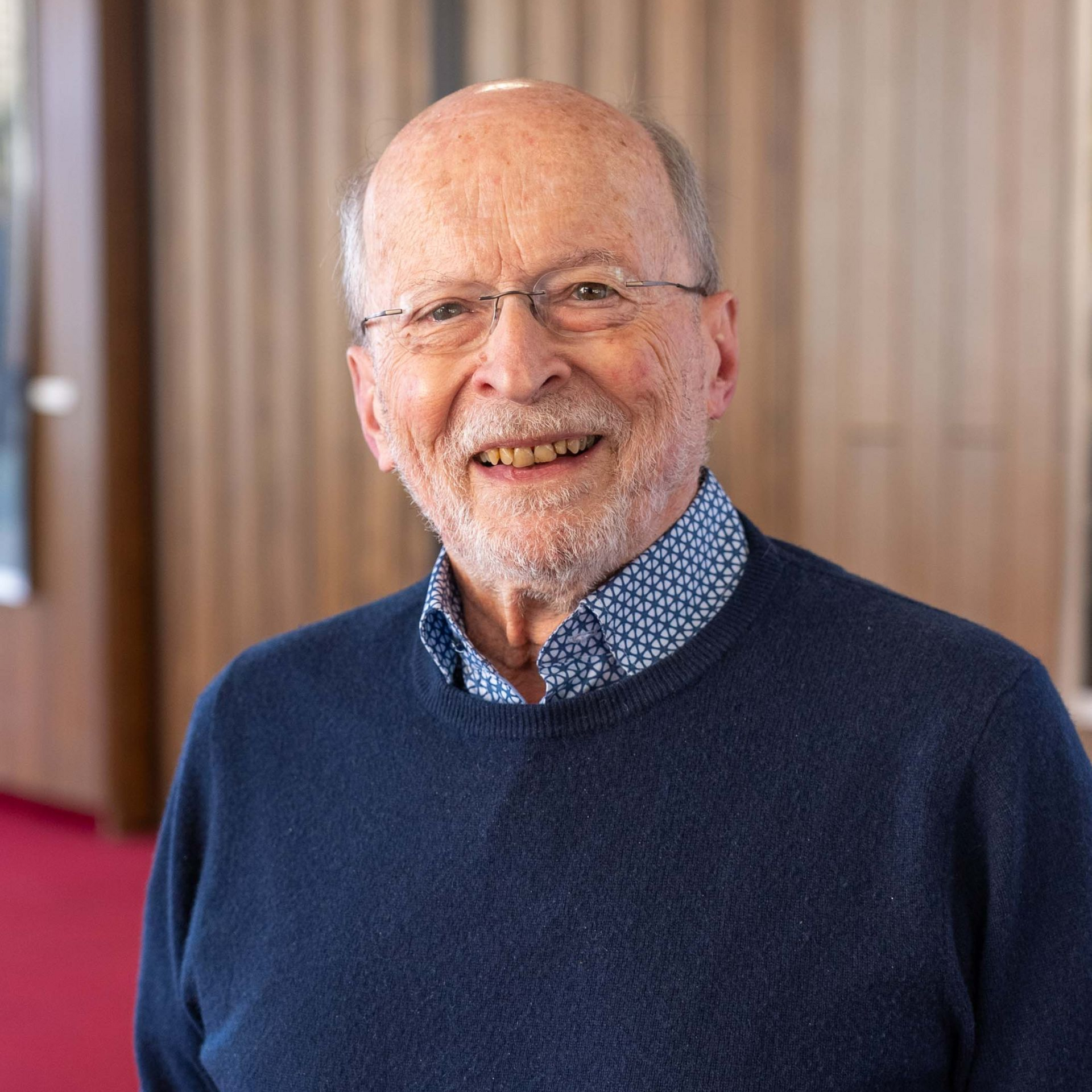 Smiling man in glasses and a blue sweater standing indoors against a wood-paneled background