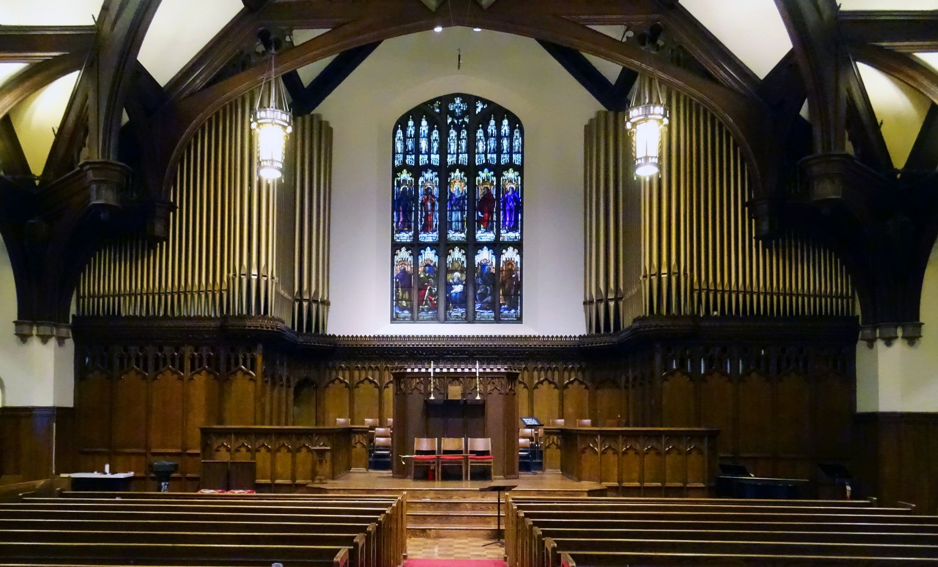 Empty church sanctuary with wooden pews, organ pipes, and a large stained-glass window above the altar.