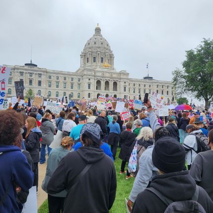 Crowd gathers on a lawn before a grand capitol building with banners and signs during a rally