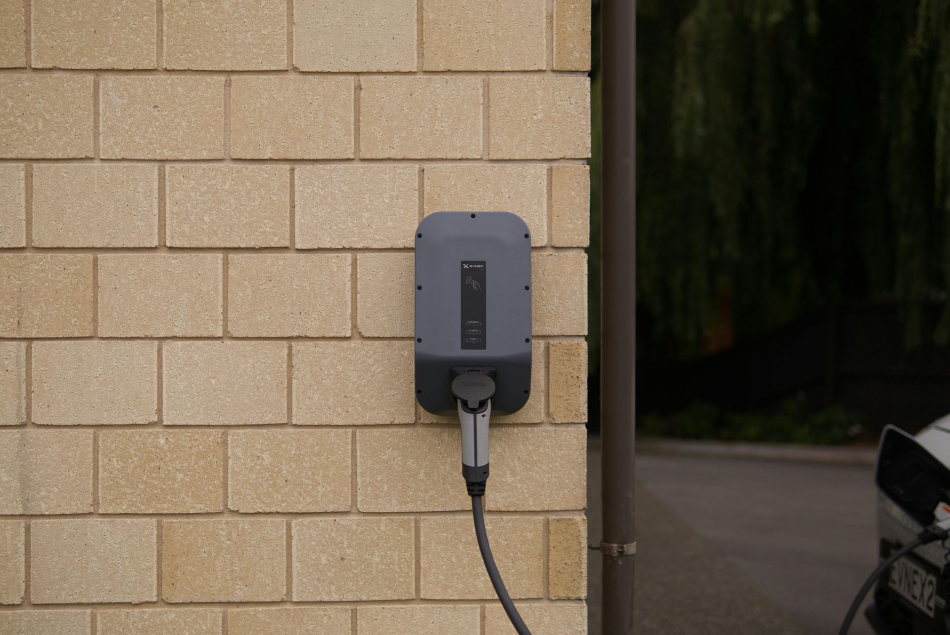 Wall-mounted gray EV charger with cable on a beige brick building exterior.