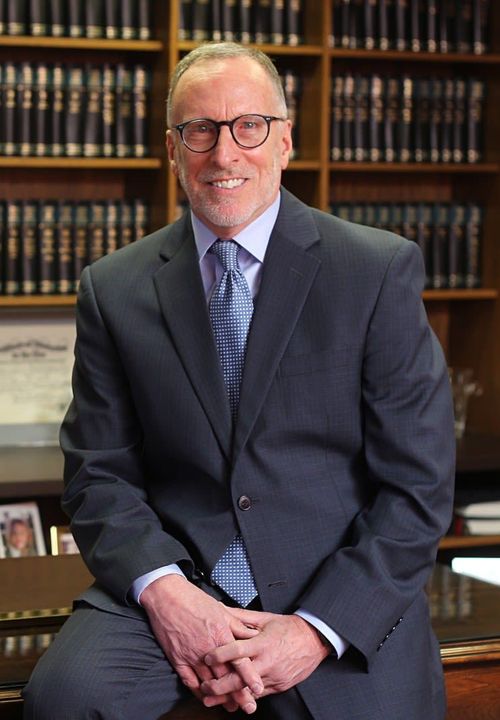 A man in a suit and tie is sitting in front of a bookshelf.