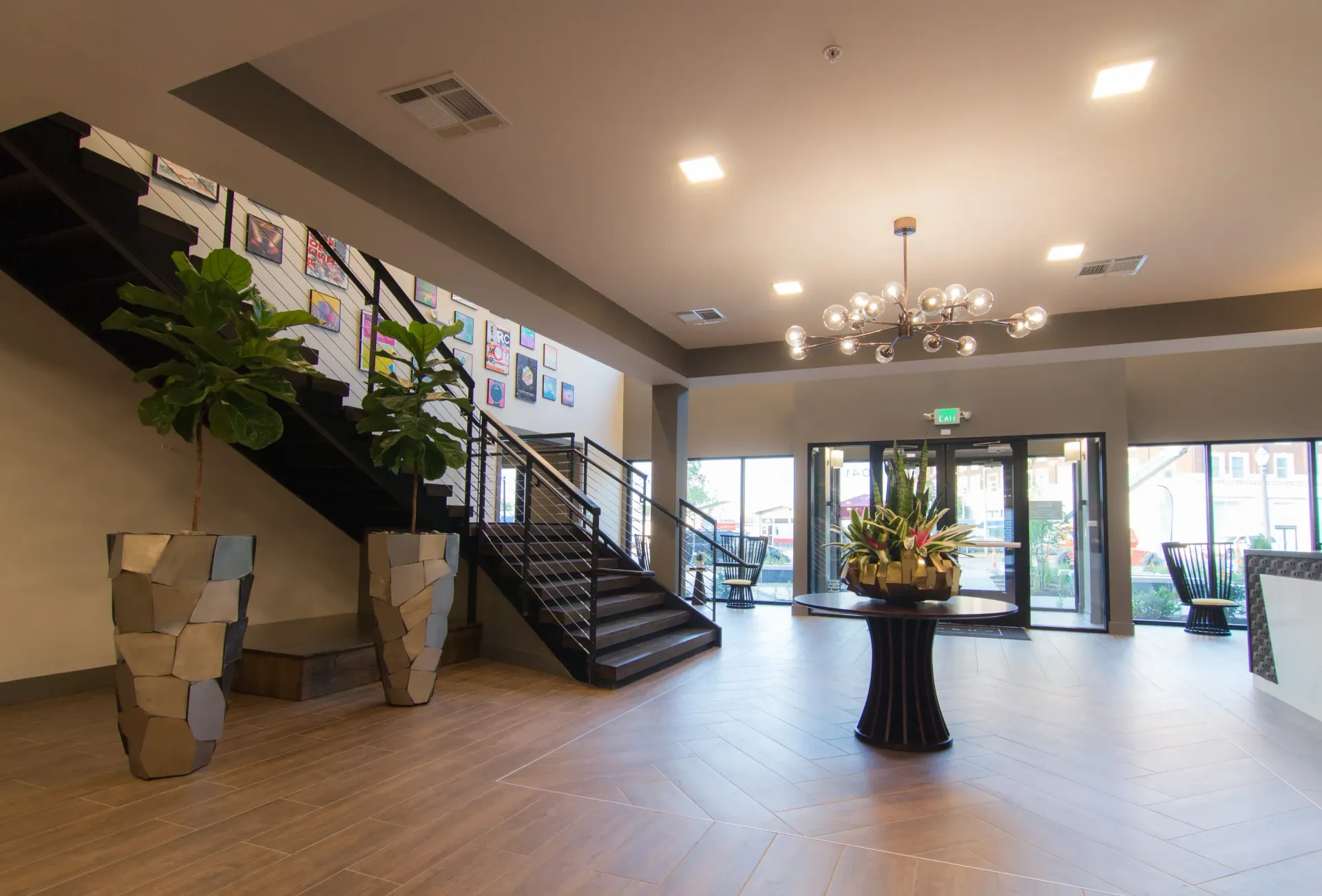 Modern apartment lobby with a staircase, decorative plants, and a round centerpiece table near glass doors.