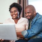 A smiling couple sits together indoors looking at a laptop screen.