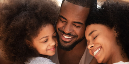 Three people with curly hair are embracing closely, smiling with their eyes closed in a warm, affectionate moment.