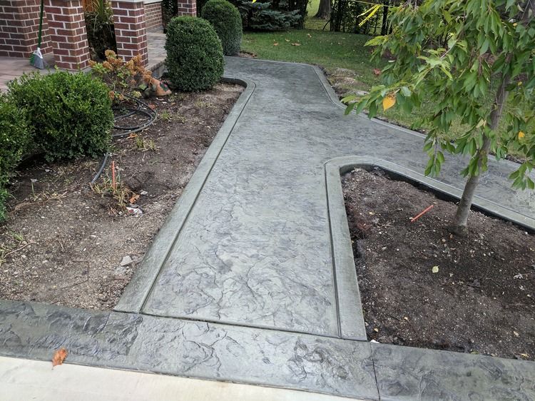 A concrete walkway leading to a house with a brick building in the background.