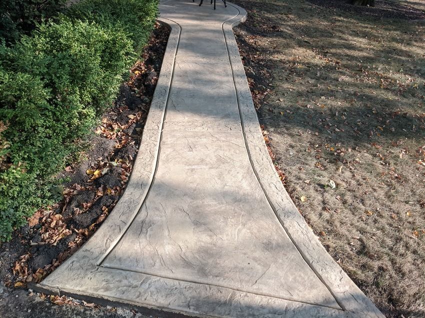 A concrete walkway with a curved design is surrounded by bushes and dirt.