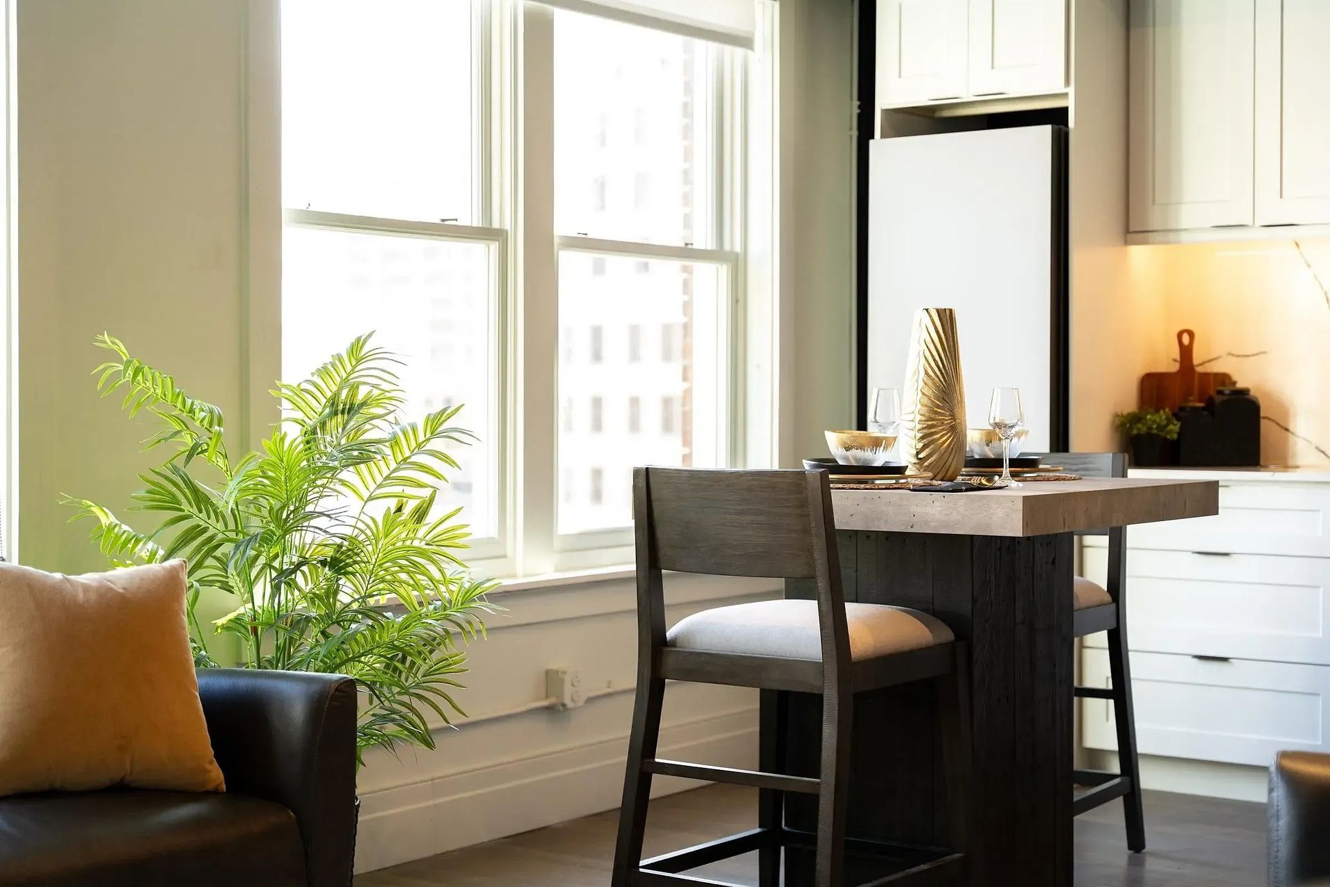 Breakfast nook with small table, two stools, and large windows. Green plant, gold vase, and brown leather chair.