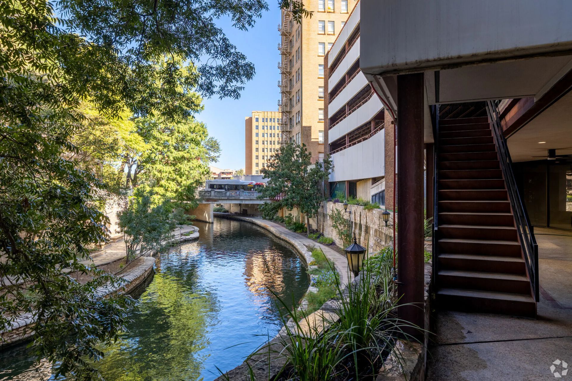 Riverwalk scene in San Antonio, Texas; stone walkway, river with reflections, bridge, buildings, staircase.