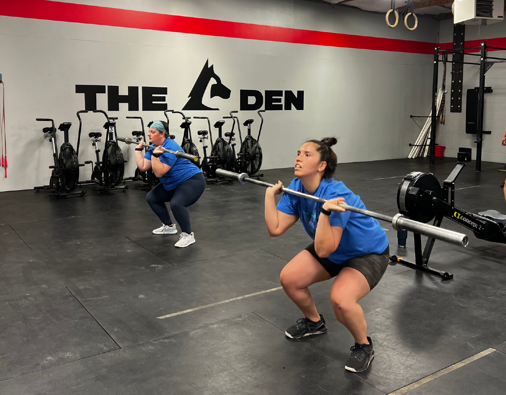 Two women are squatting with barbells in a gym.