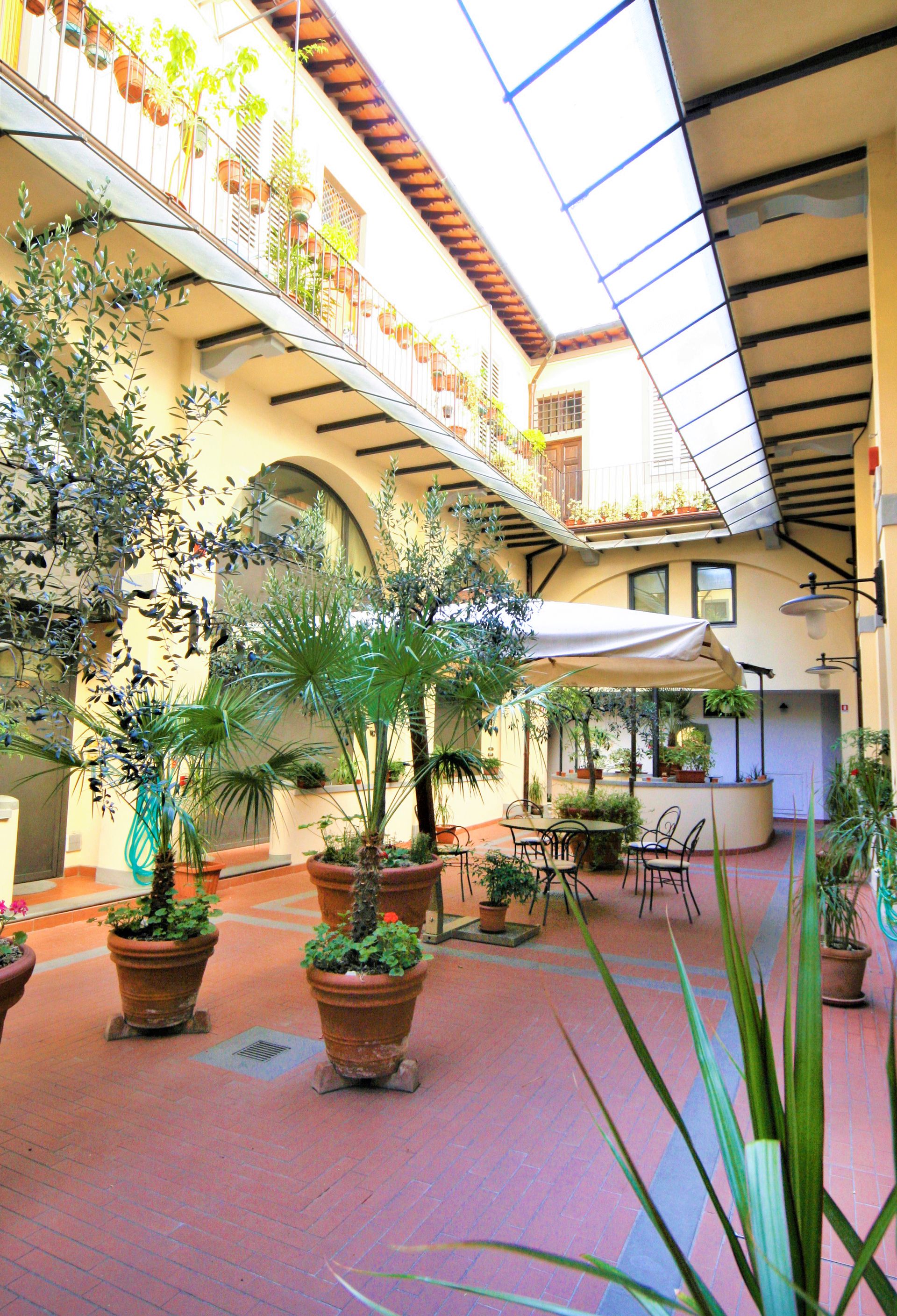 Sunlit courtyard with potted plants, arched balconies, and a small kiosk beneath a glass roof.