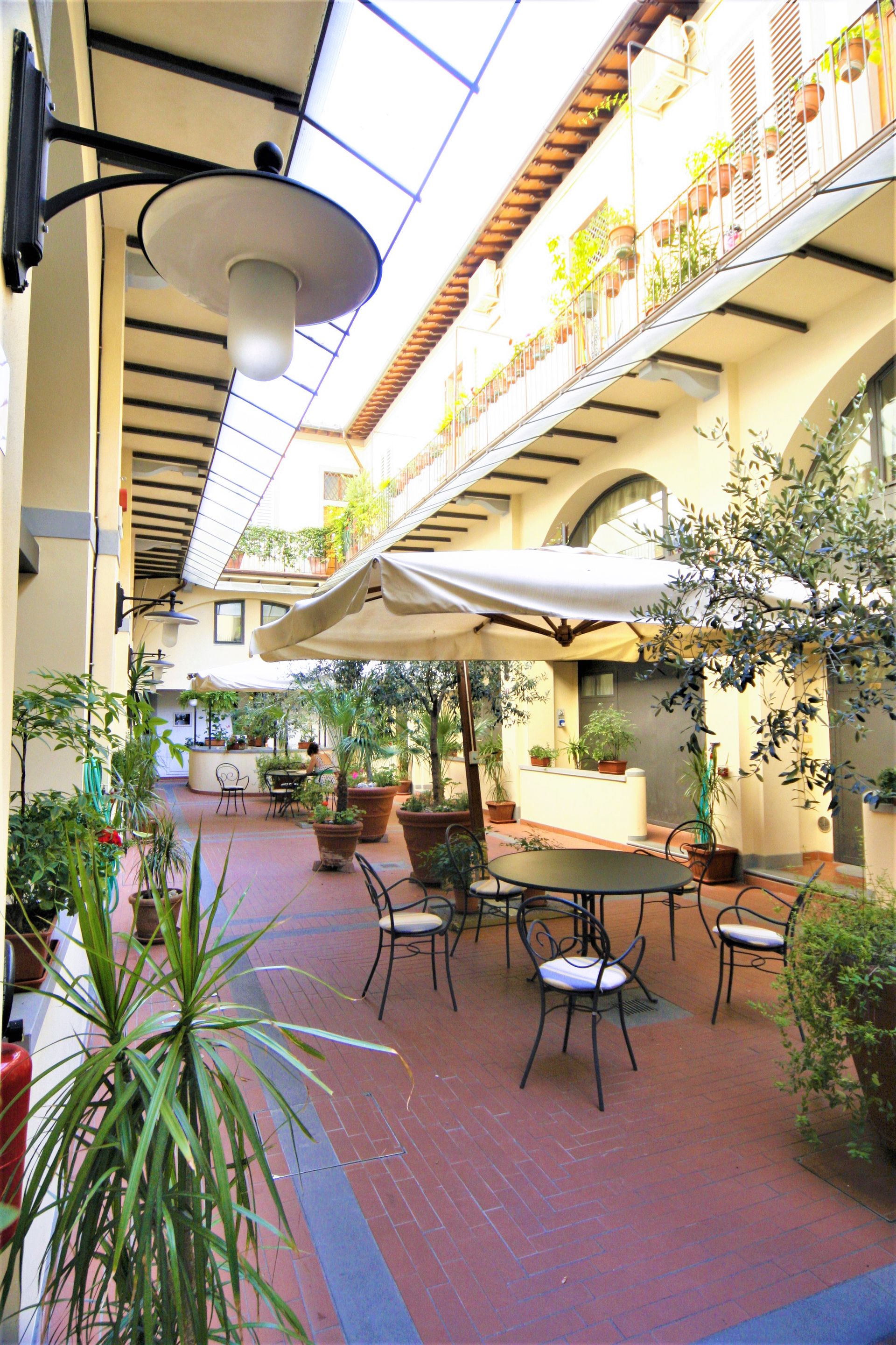 Outdoor café courtyard with red tile floor, white umbrellas, and potted plants along a sunlit building corridor