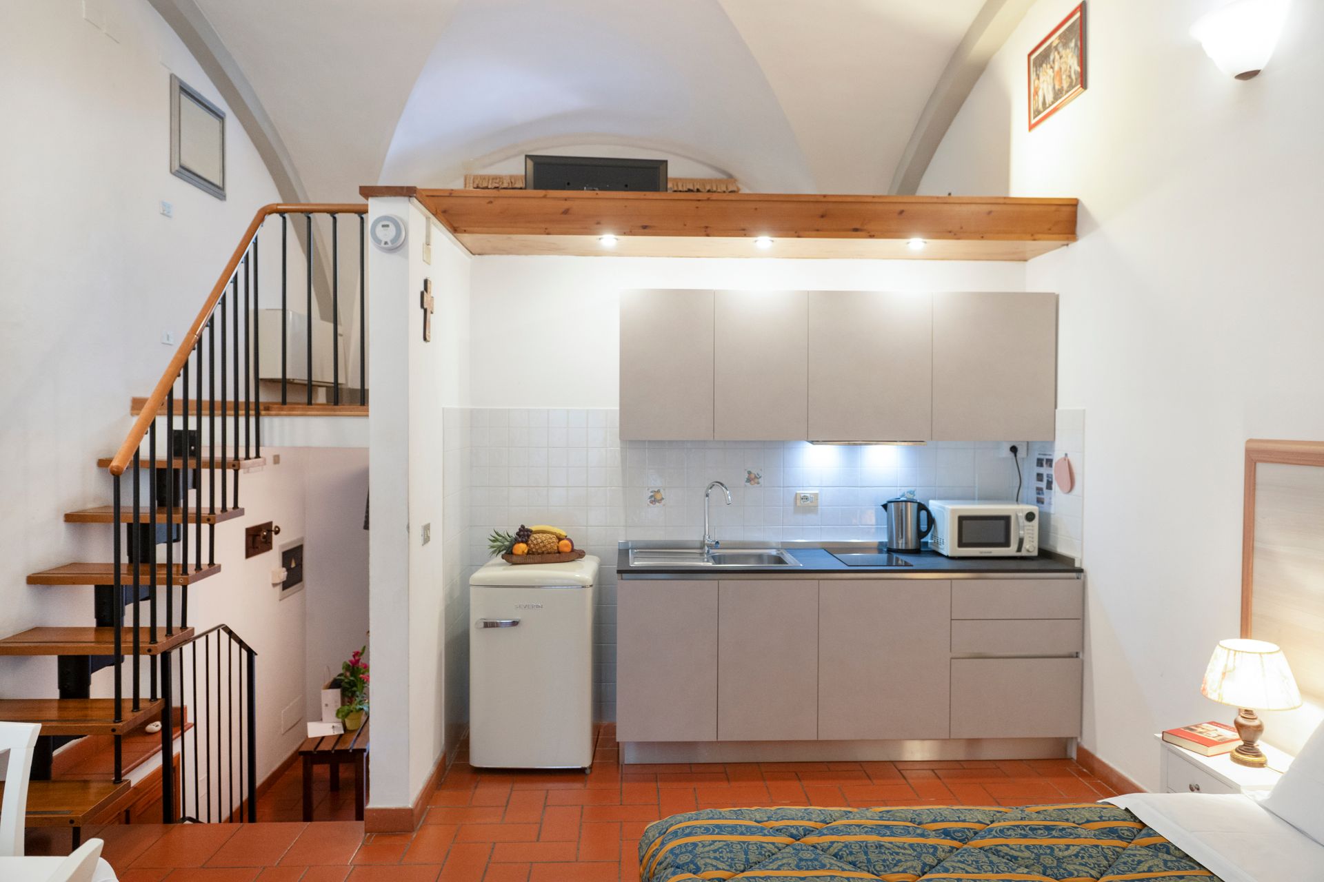 Loft studio apartment with kitchen, staircase, and bed under a high arched ceiling