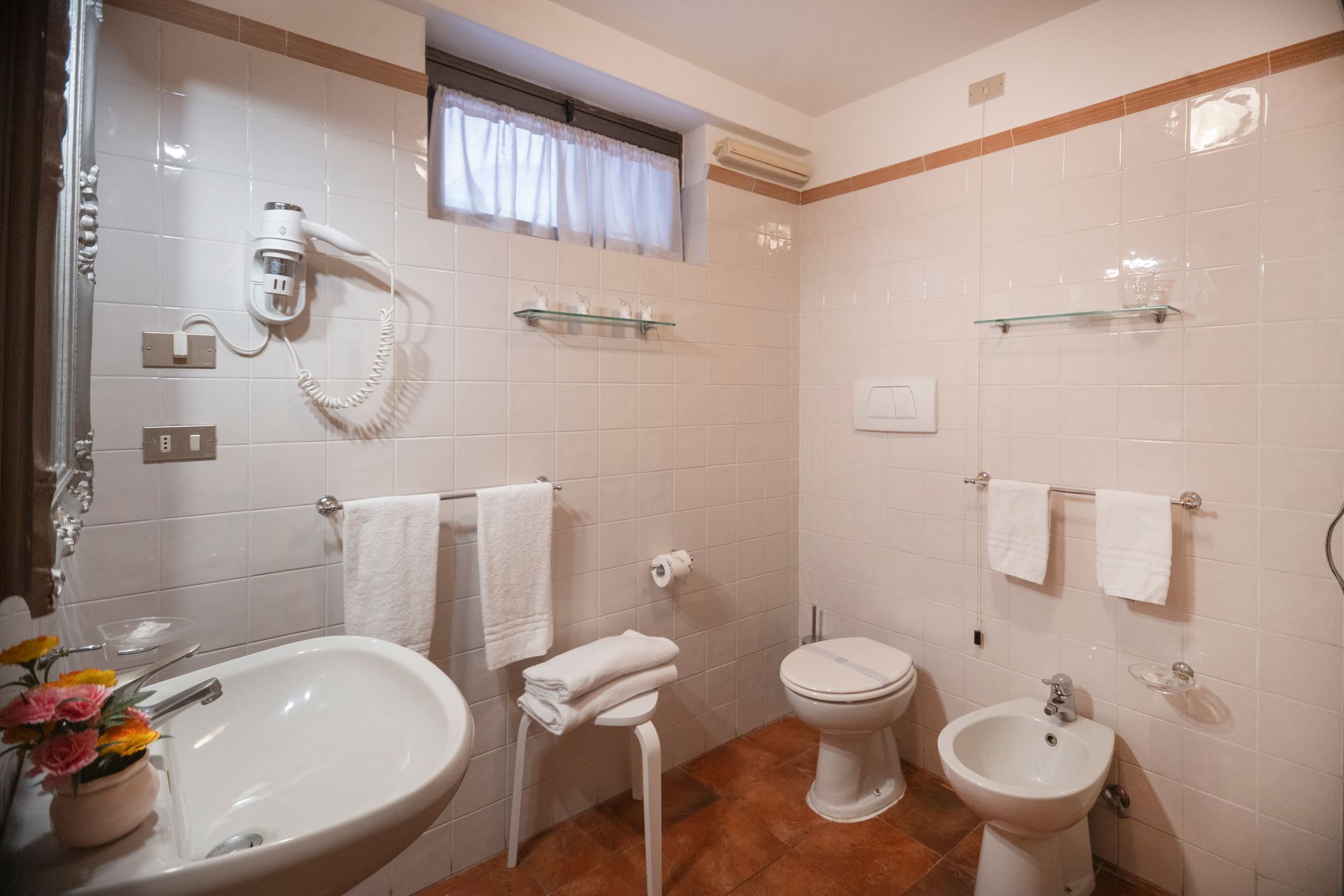 Bright bathroom with beige tiles, toilet, bidet, sink, towels, and a small window.