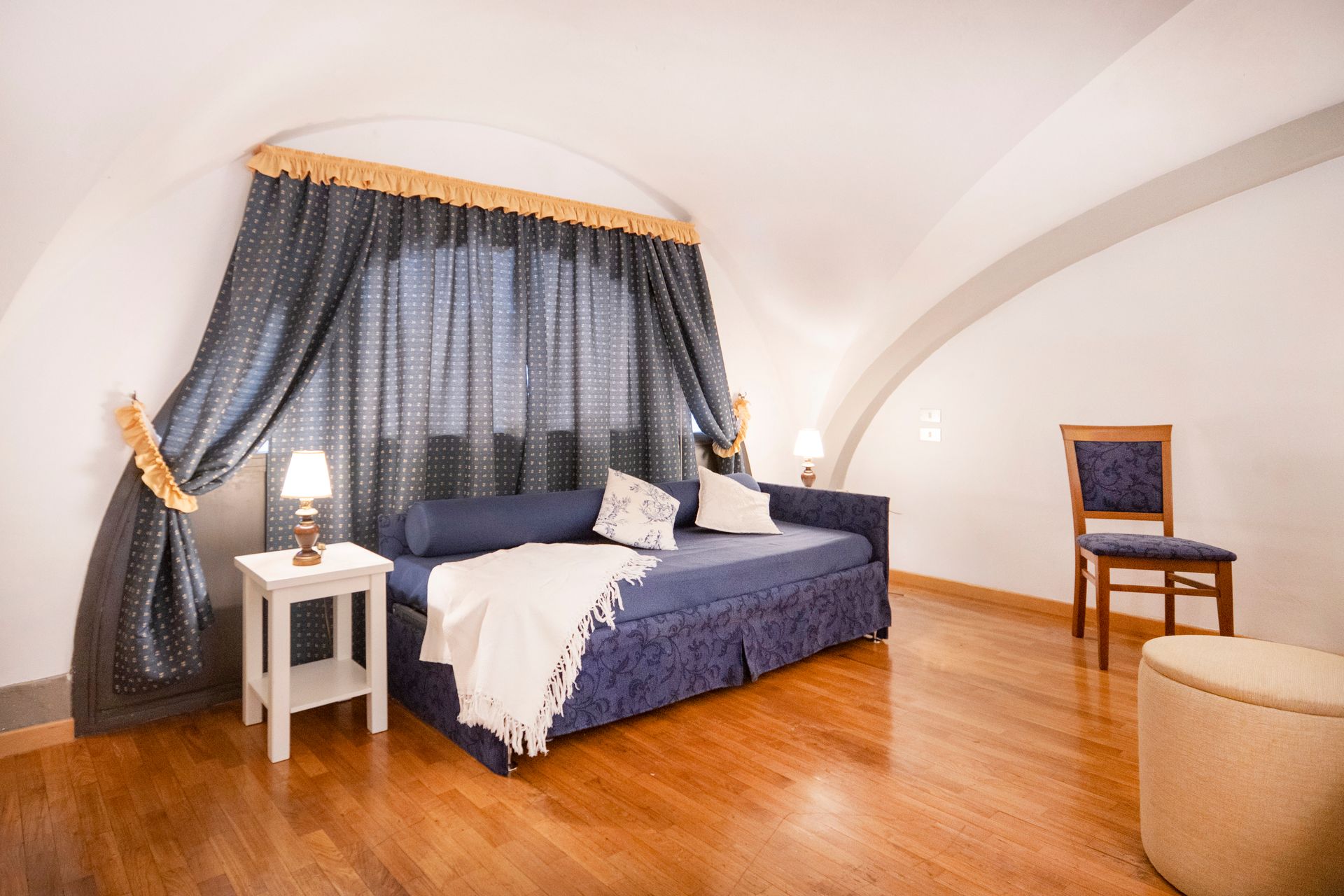 Cozy bedroom with a blue bed, dark curtains, wood floor, and a small chair by the wall