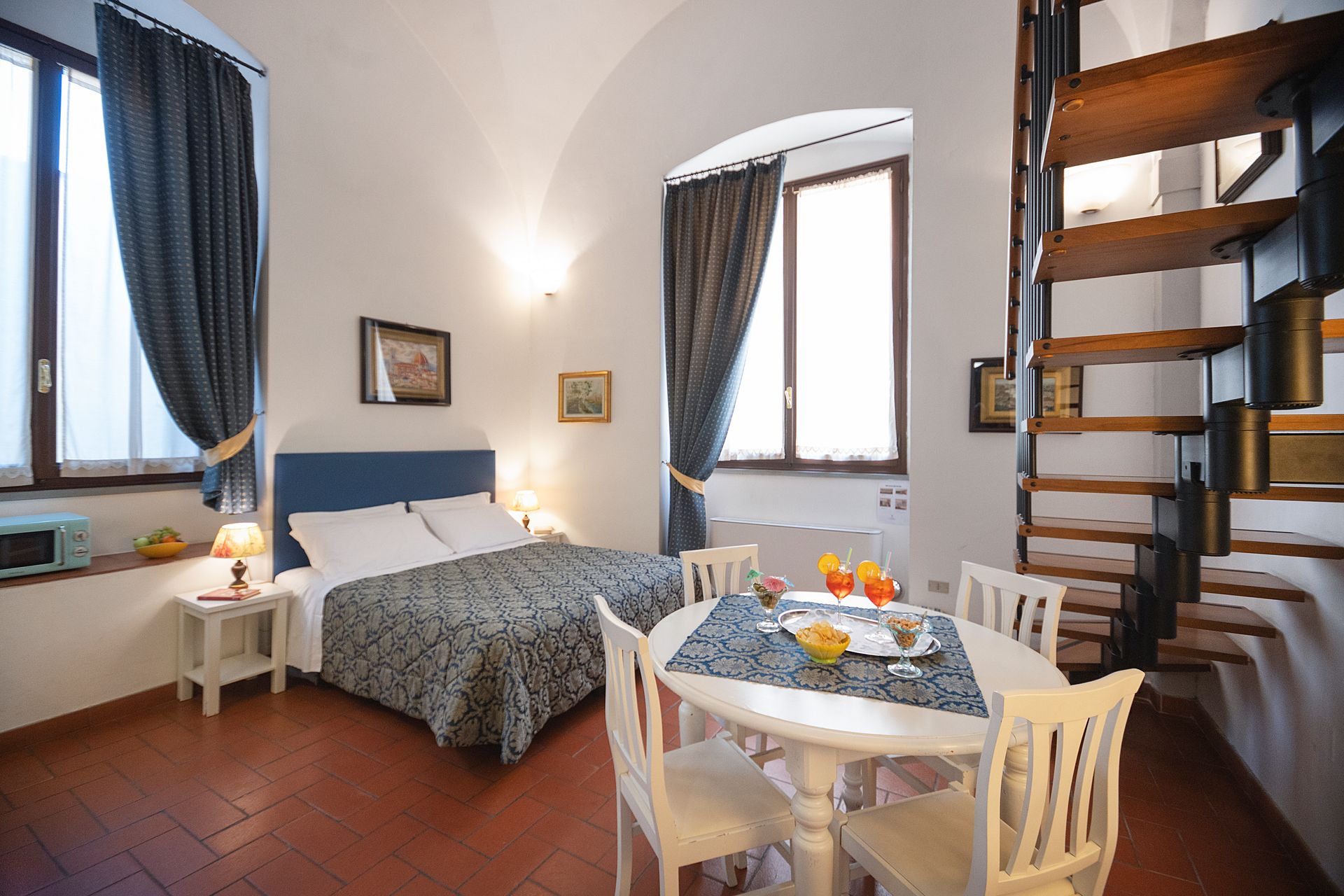 Bright bedroom with double bed, small dining table, tall windows, and a wooden spiral staircase