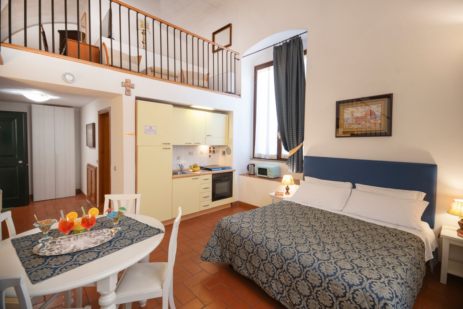 Bright bedroom with loft railing, double bed, small dining table, kitchen nook, and window with curtains