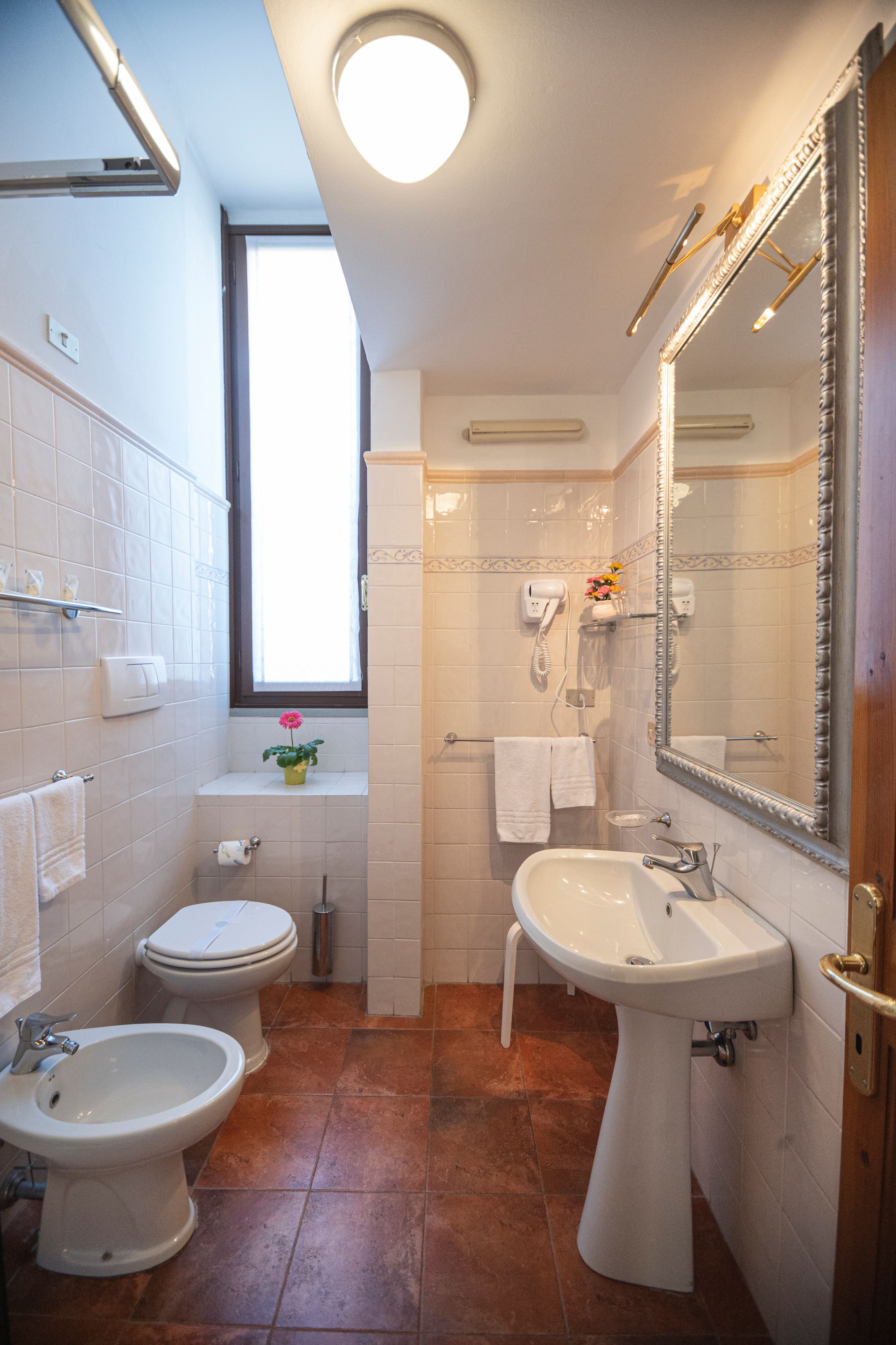 Narrow white bathroom with a sink, bidet, toilet, shower, and large mirror on terracotta tile flooring