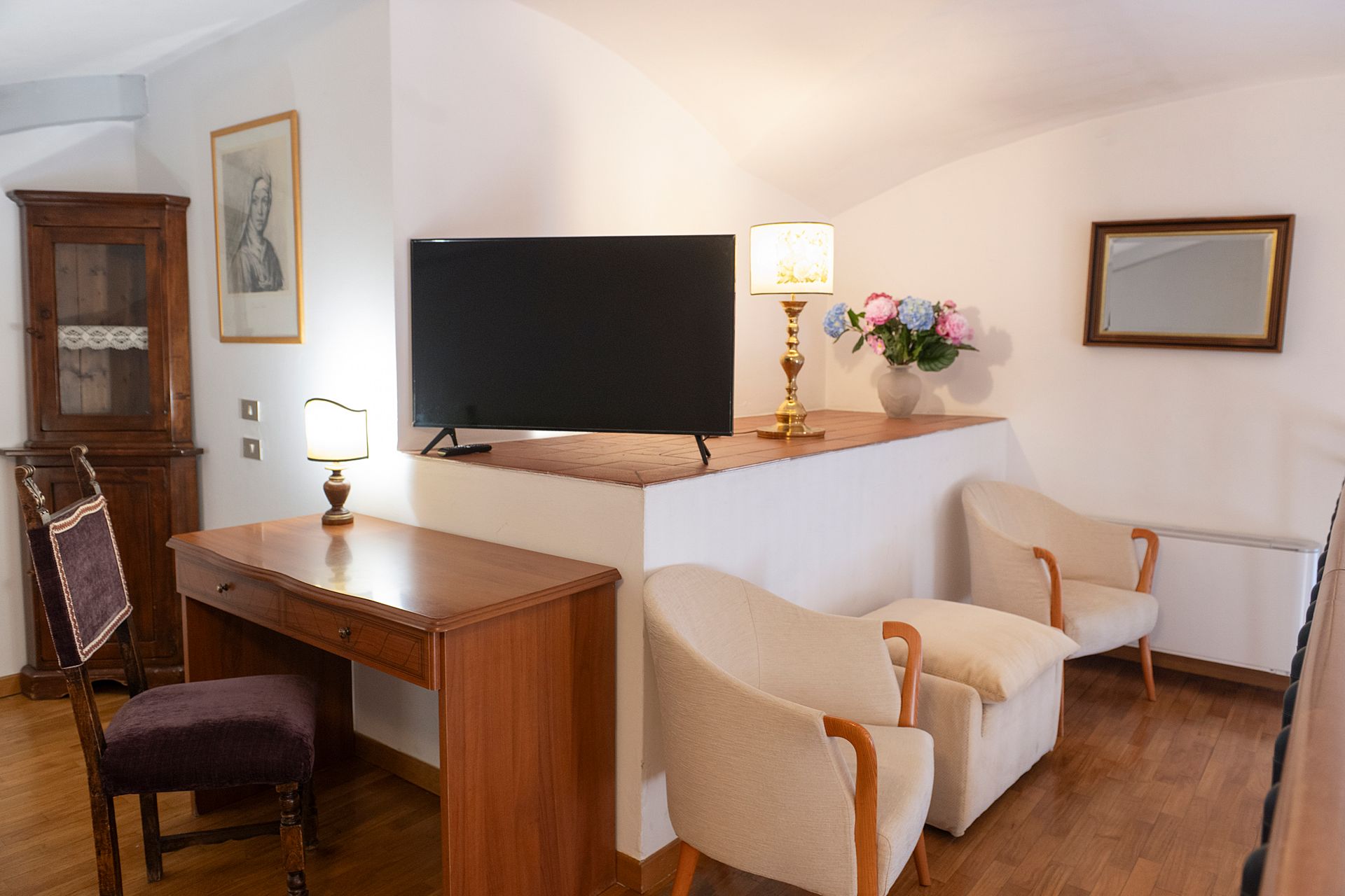 Cozy living room with cream sofa, wooden desk, lamp, flowers, wall art, and TV on a white wall
