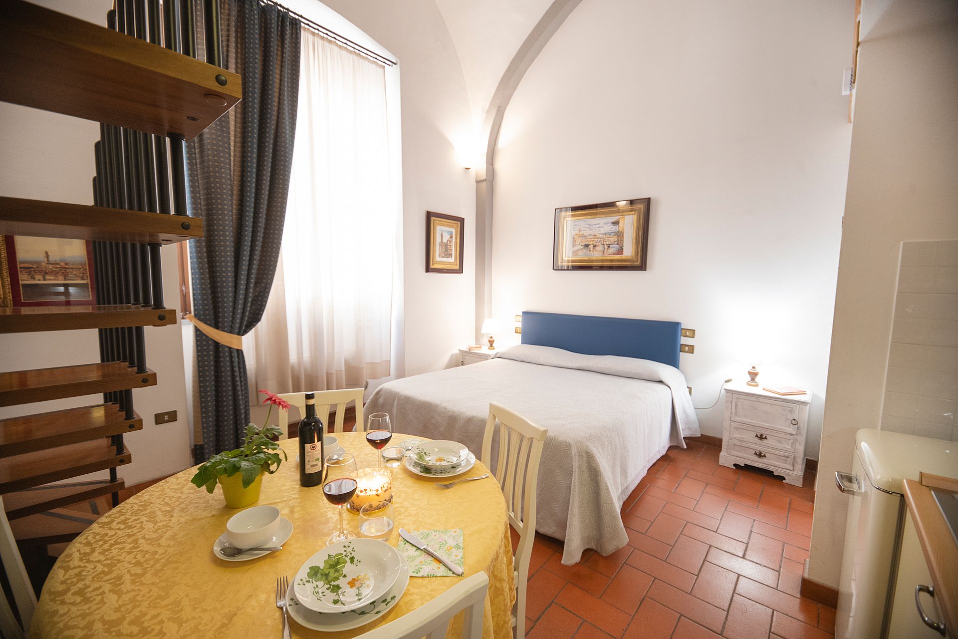 Bright bedroom with a white bed, terracotta tile floor, and a small table set for breakfast by the window.