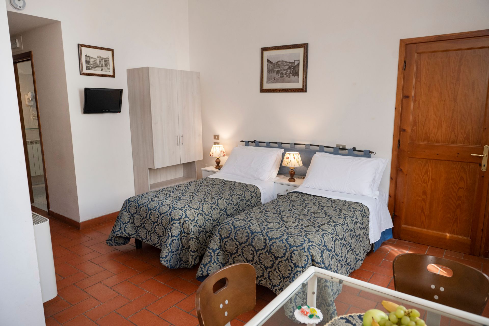 Hotel room with two twin beds, wooden wardrobe, bedside lamps, and terracotta tile floor