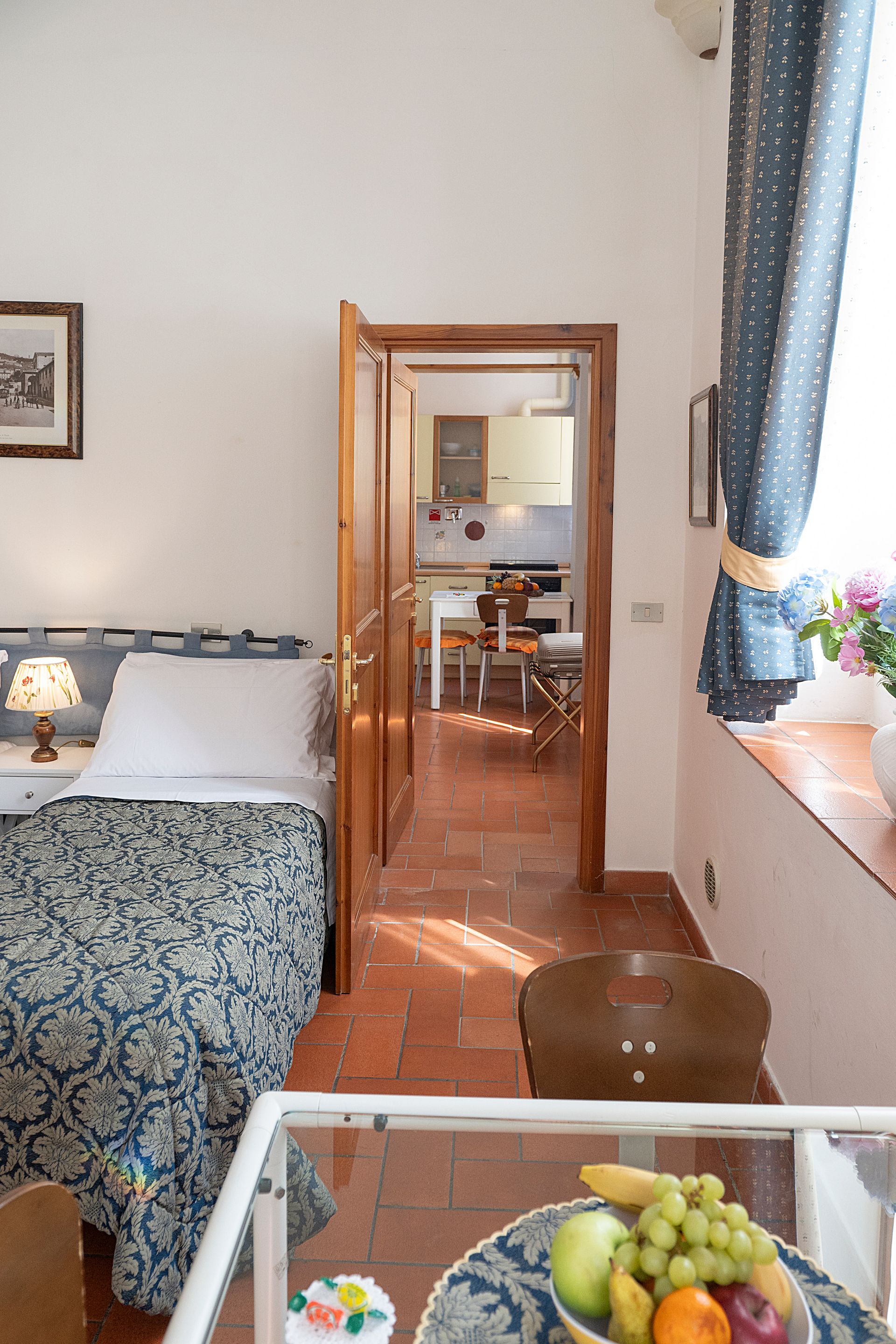 Bedroom with bed and lamp, looking through an open doorway into a dining area, with grapes on a table in front.