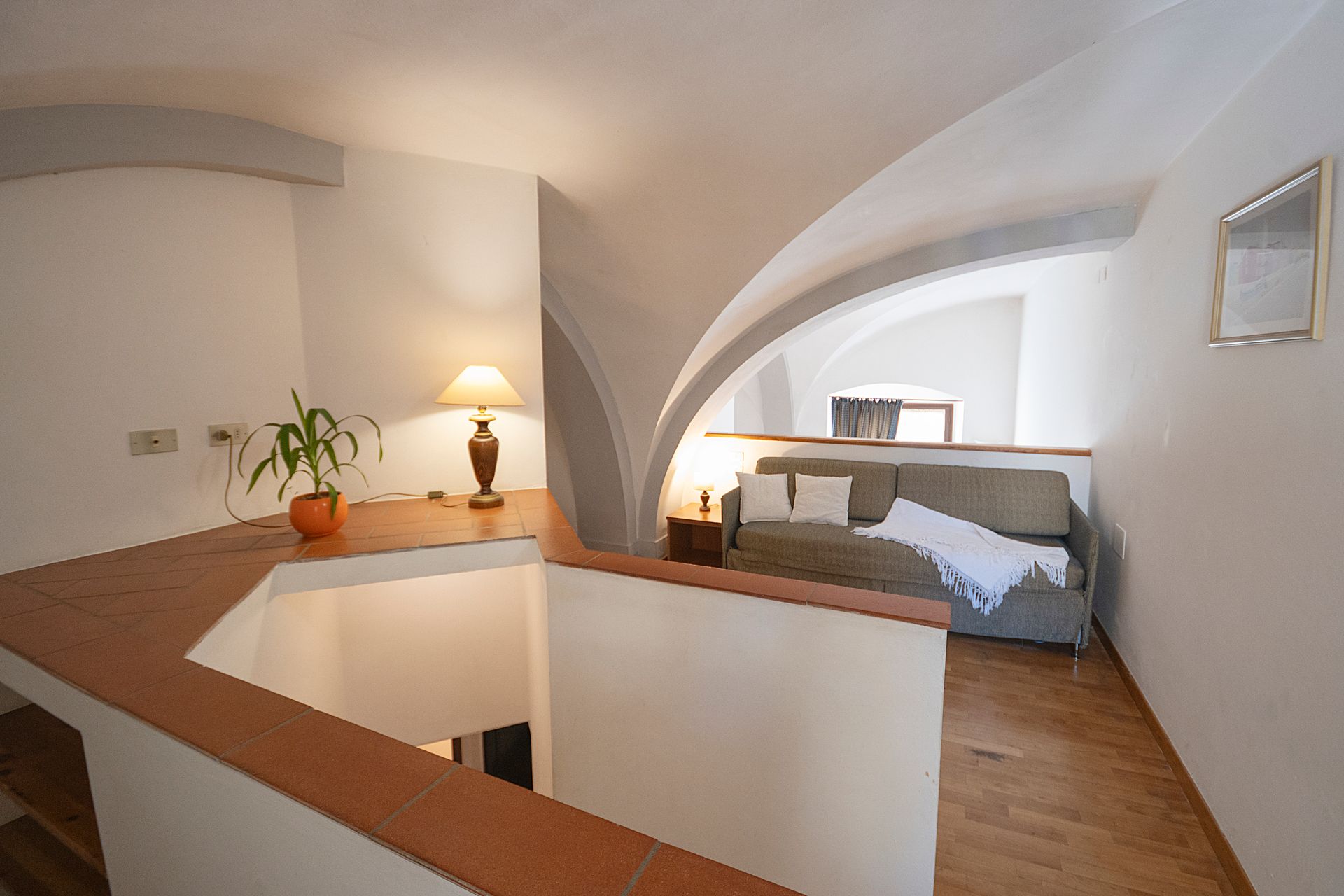 Loft bedroom with arched ceiling, wooden floors, bed, lamp, and a small desk by the railing