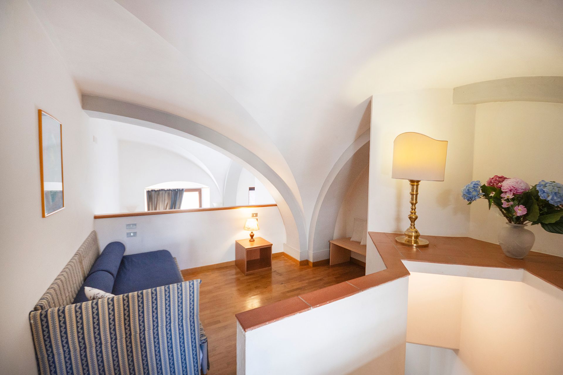 Bright white living area with arched ceilings, blue sofa, wooden floor, and a lamp beside flowers.