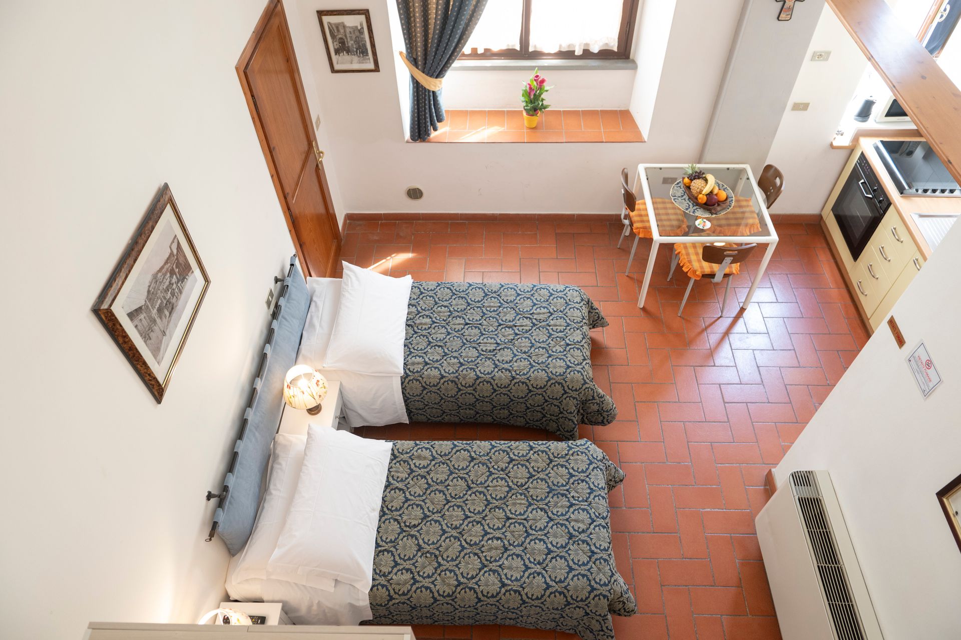 Small hotel room with two single beds, window, desk, and terracotta tile floor