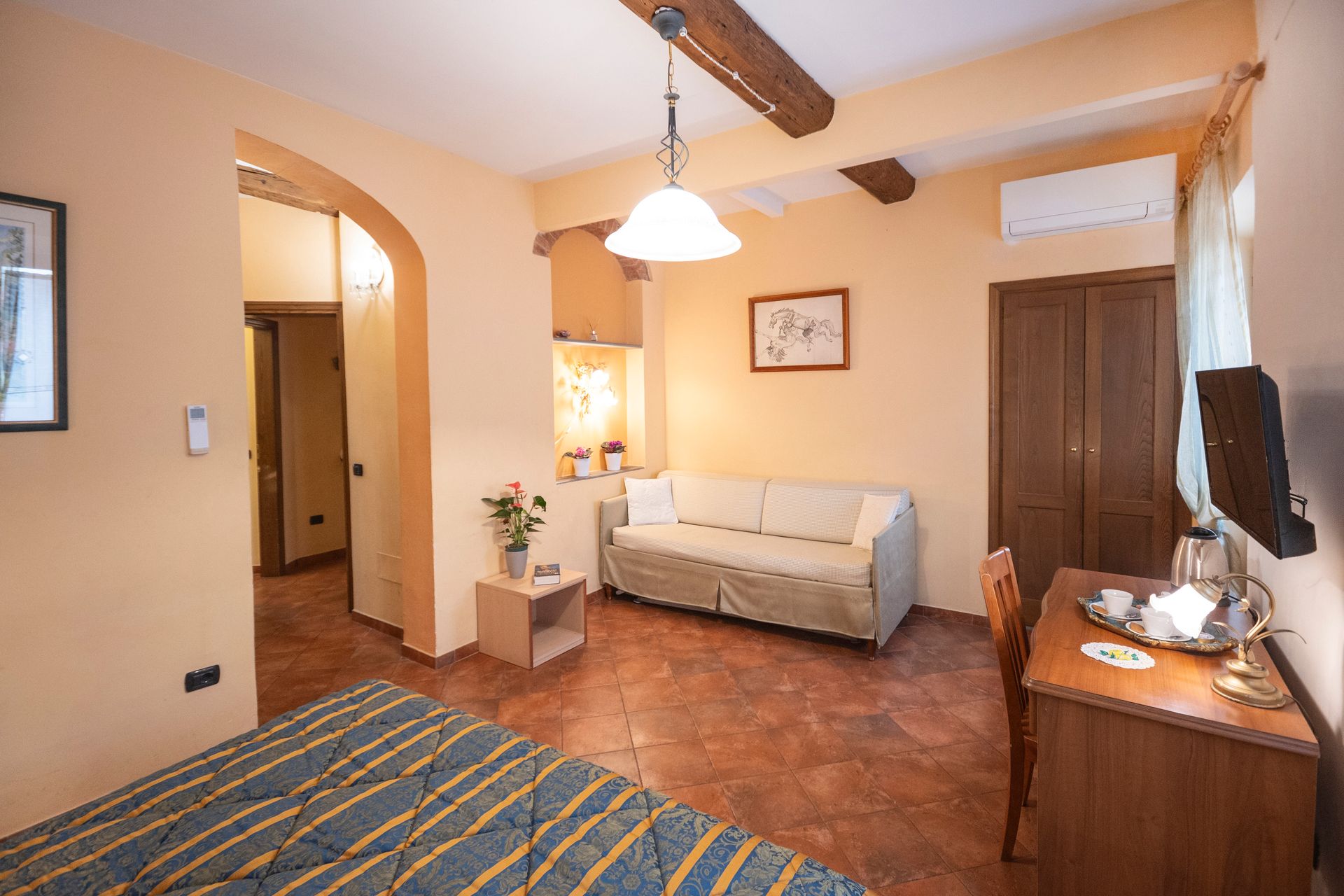 Cozy hotel room with bed, sofa, desk, terracotta tile floor, and ceiling fan in warm light