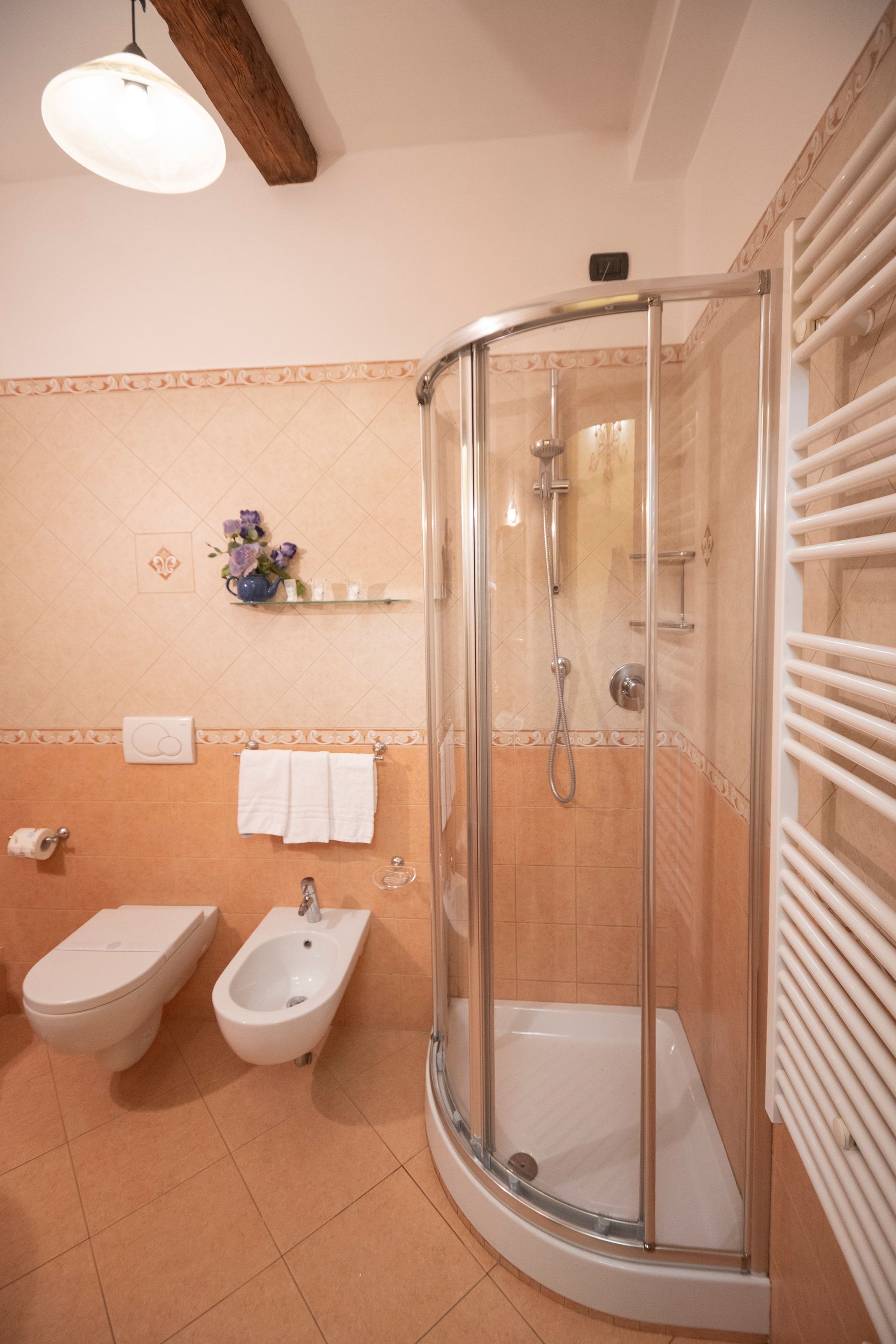 Small beige bathroom with wall-mounted toilet, bidet, and a curved glass shower enclosure.