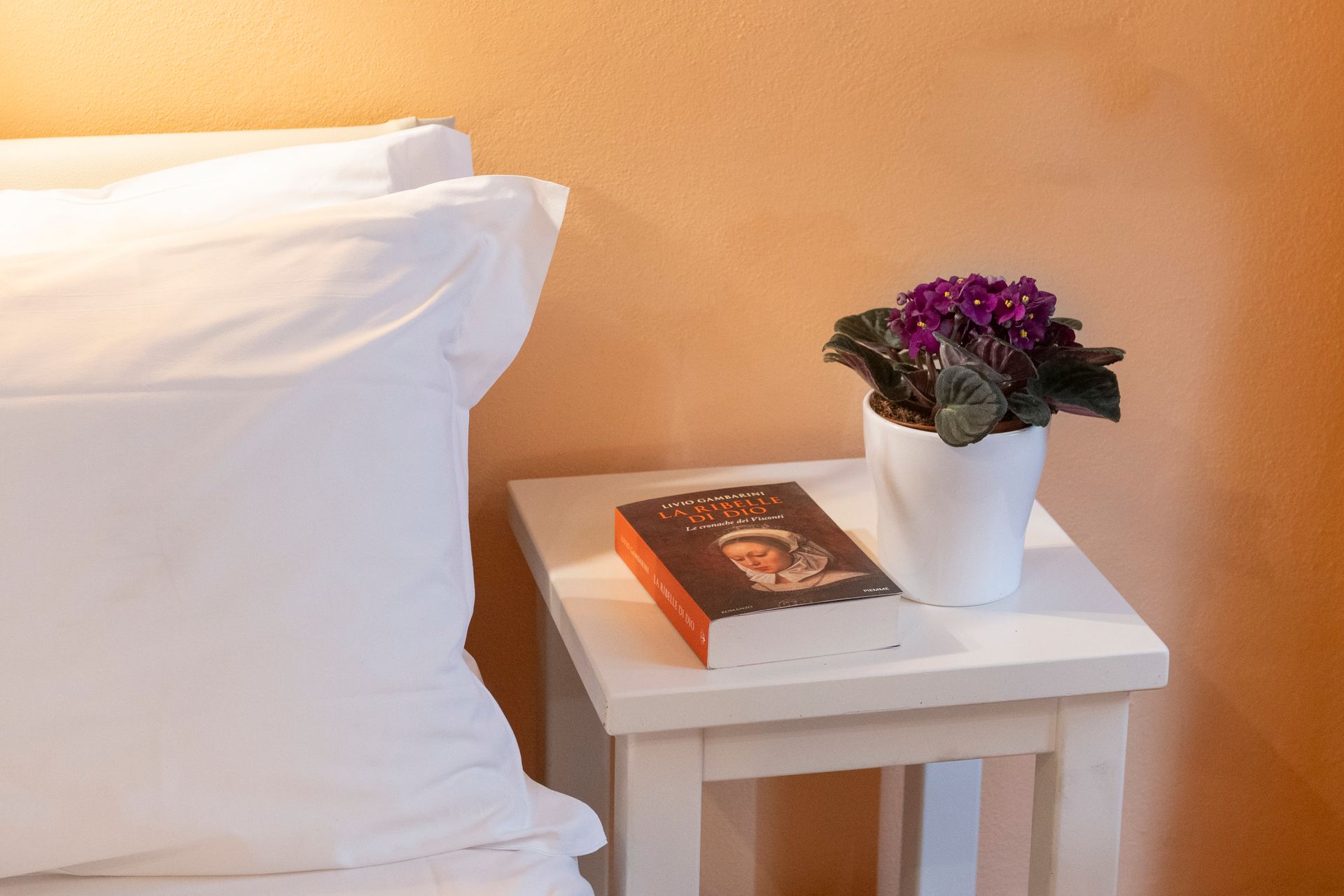 White bed beside a small table with a book and purple flowers in a white pot against a tan wall
