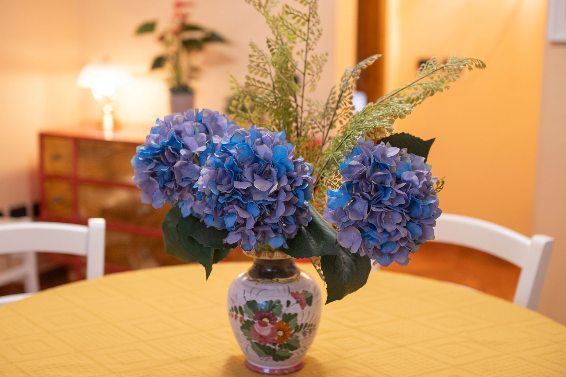 Blue hydrangeas in a floral vase on a yellow table in a warm dining room