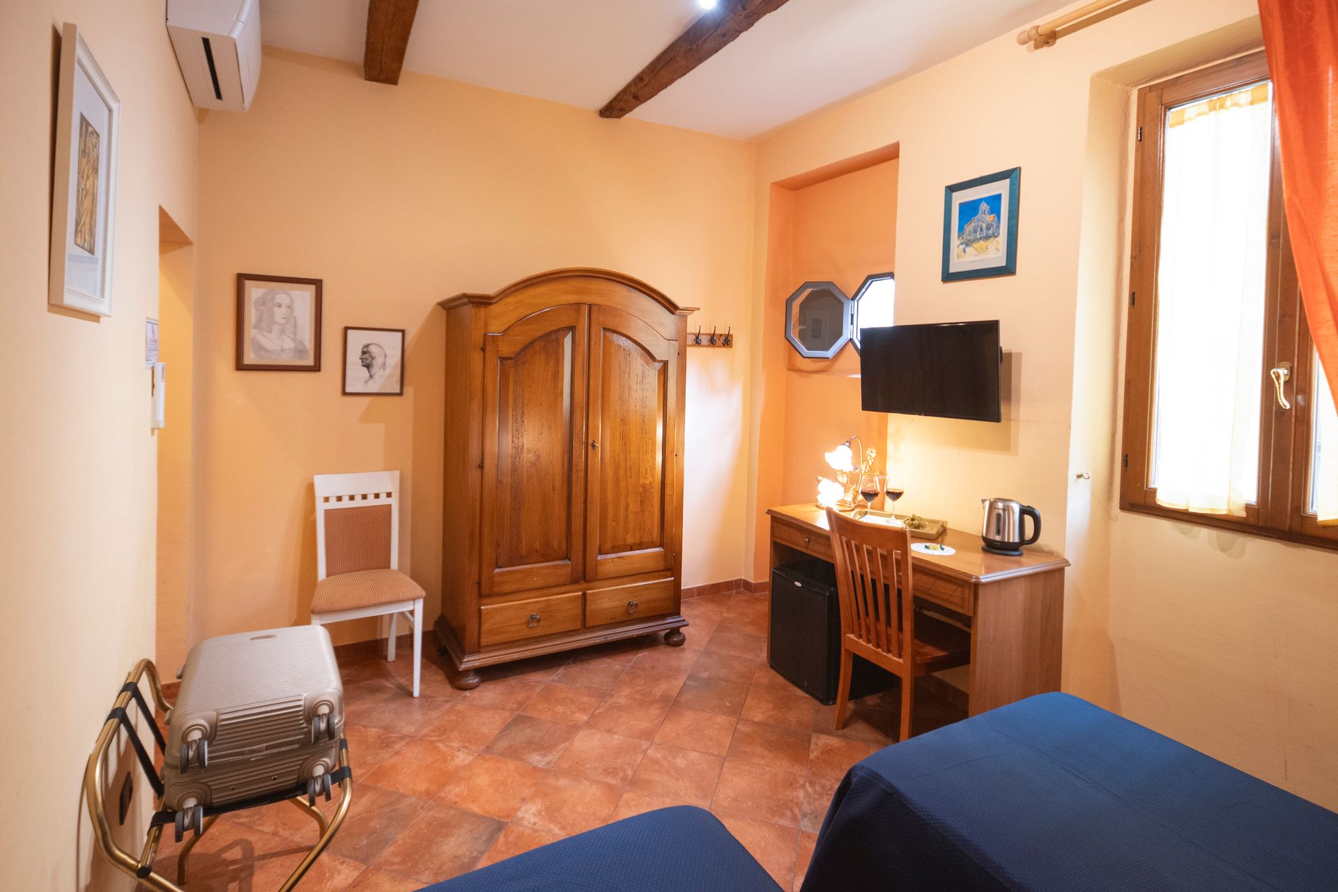 Small cozy bedroom with wooden wardrobe, desk, wall-mounted TV, and blue bedspread