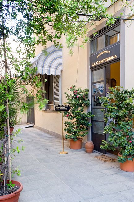 Narrow patio walkway beside a café entrance with potted plants and leafy trees