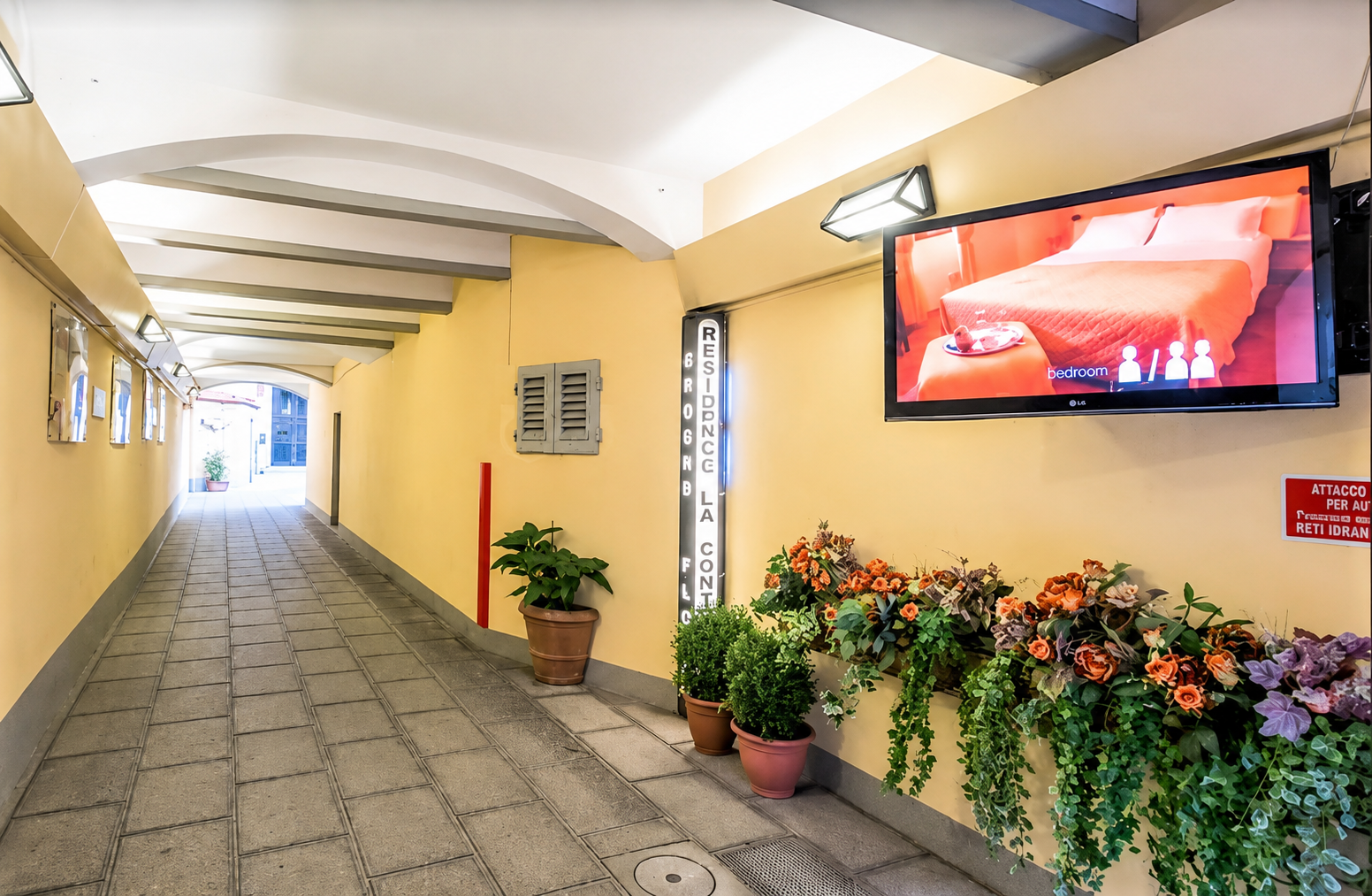 Bright indoor corridor with potted flowers, a wall-mounted TV, and patterned tiled floor