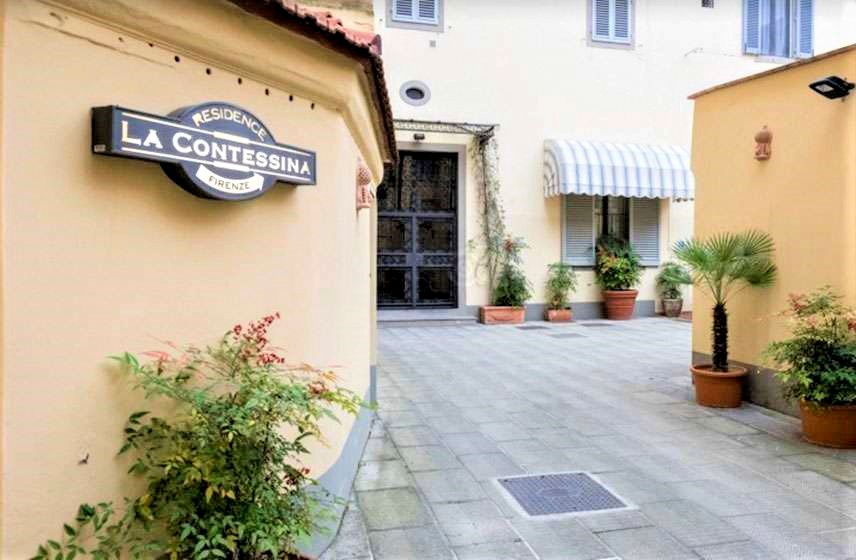 Narrow courtyard entrance with a “La Conseria” sign, potted plants, and pale yellow stucco walls