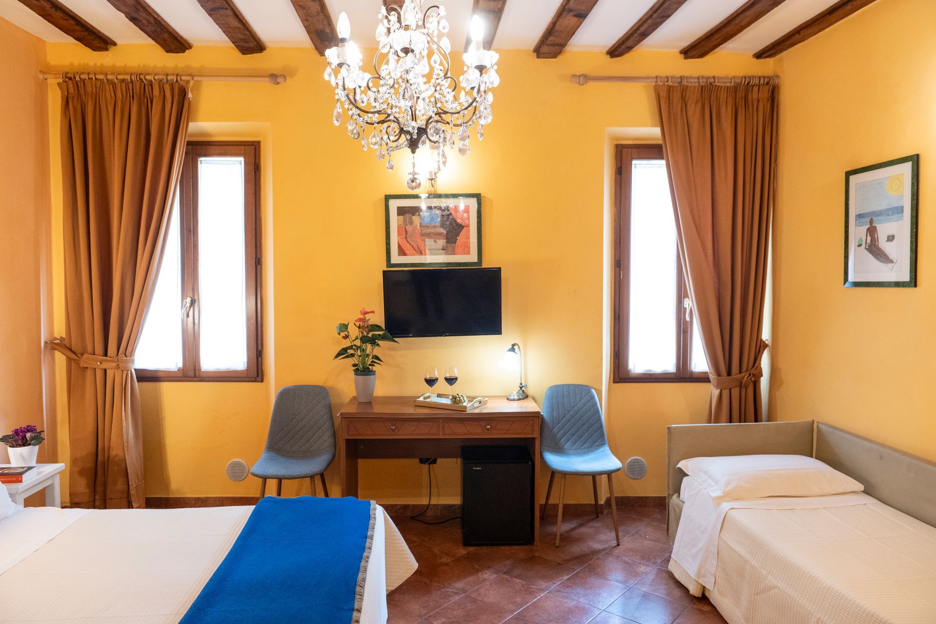 Bright hotel room with two beds, desk, blue chairs, and yellow walls under a chandelier