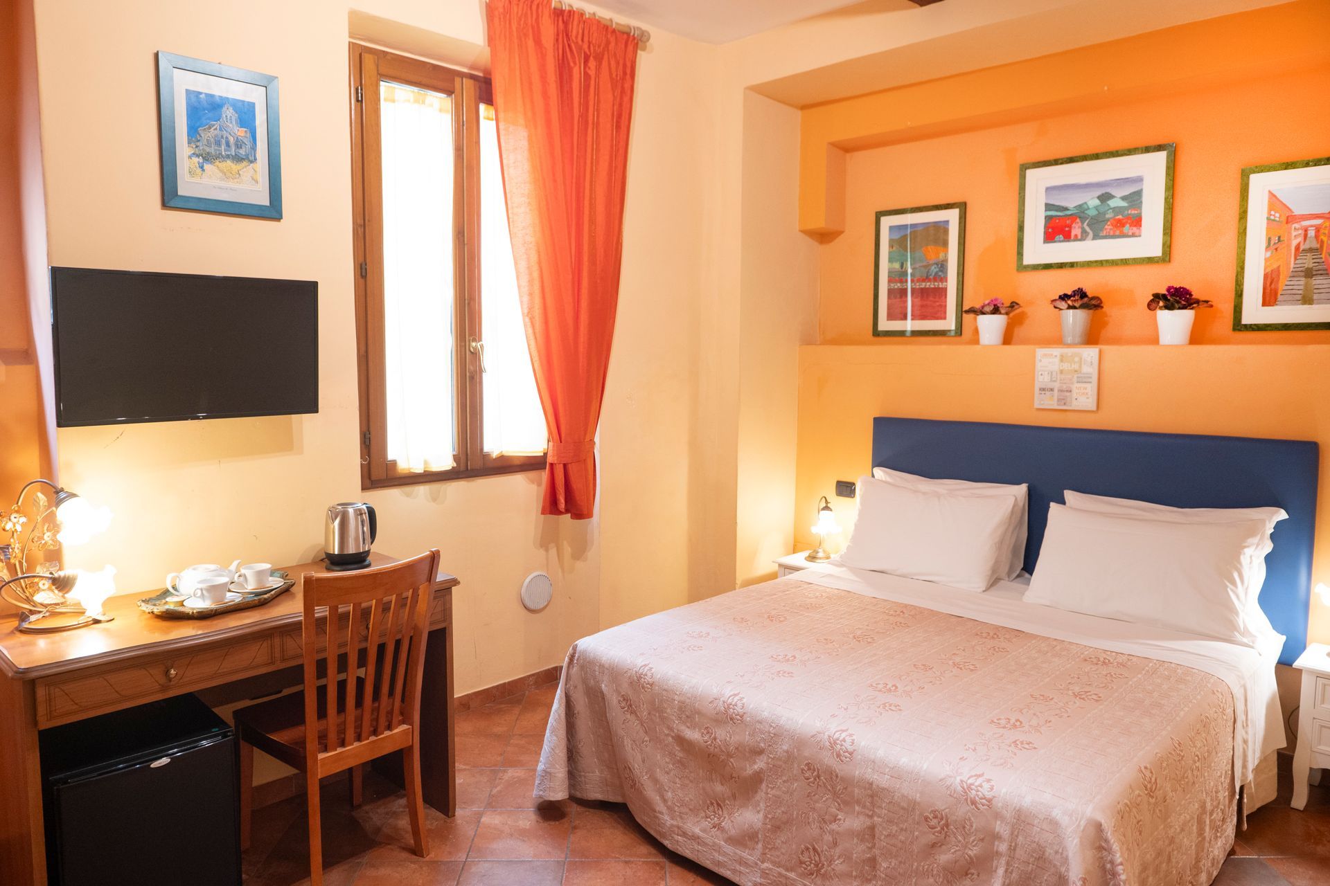 Cozy hotel room with a bed, desk, wall-mounted TV, window, and orange curtains