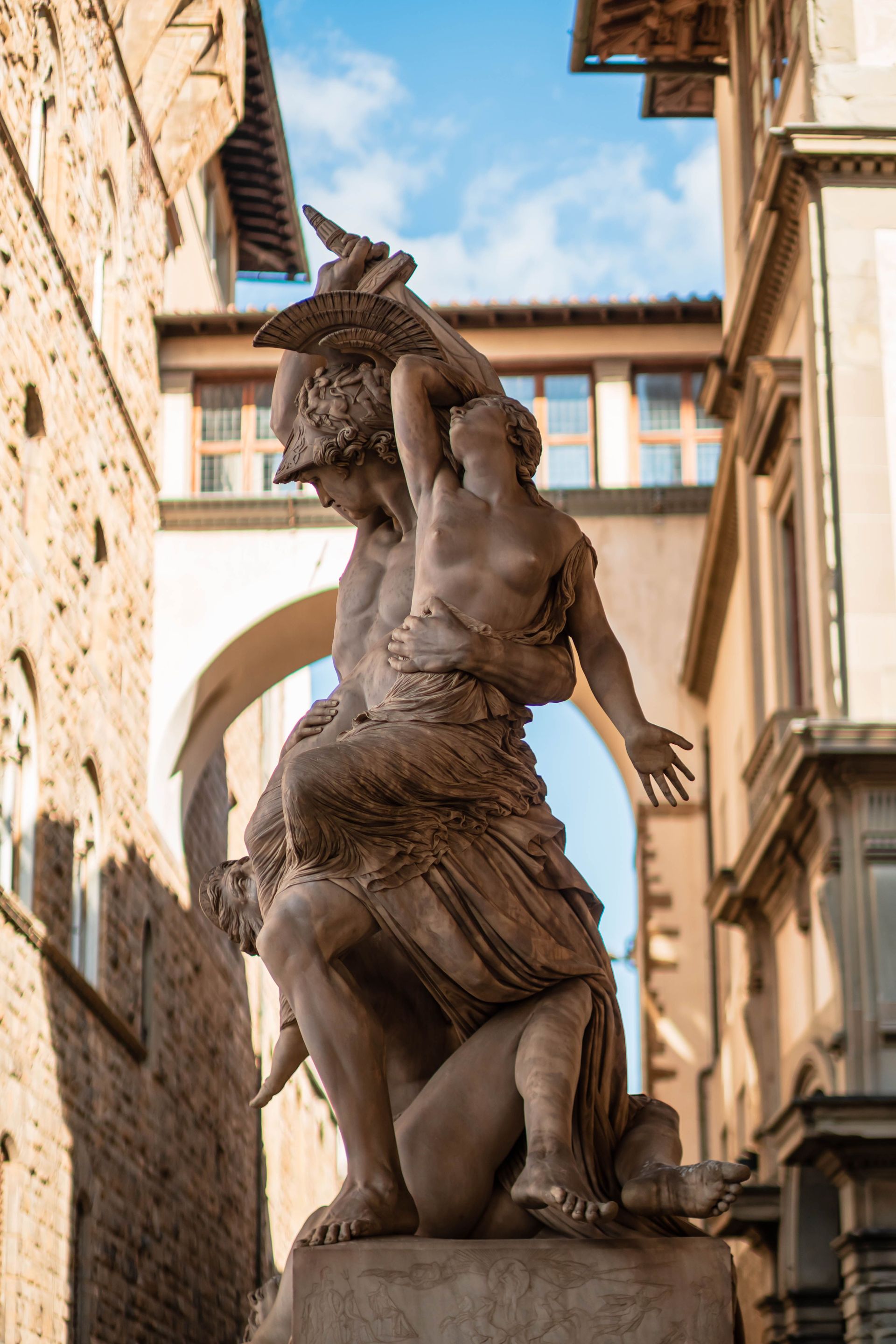 Stone statue in a narrow European street, with a raised arm holding an object beneath an archway