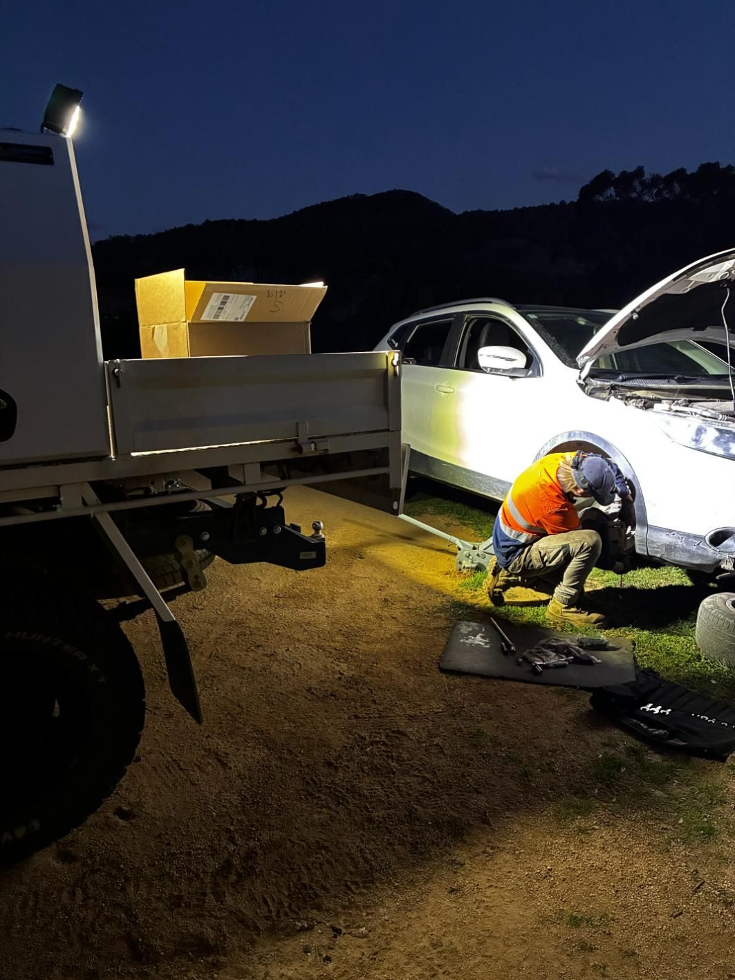Person working on a white car under a spotlight at night near a truck, mountain in background. — Bodes All Mechanical In Merimbula, NSW