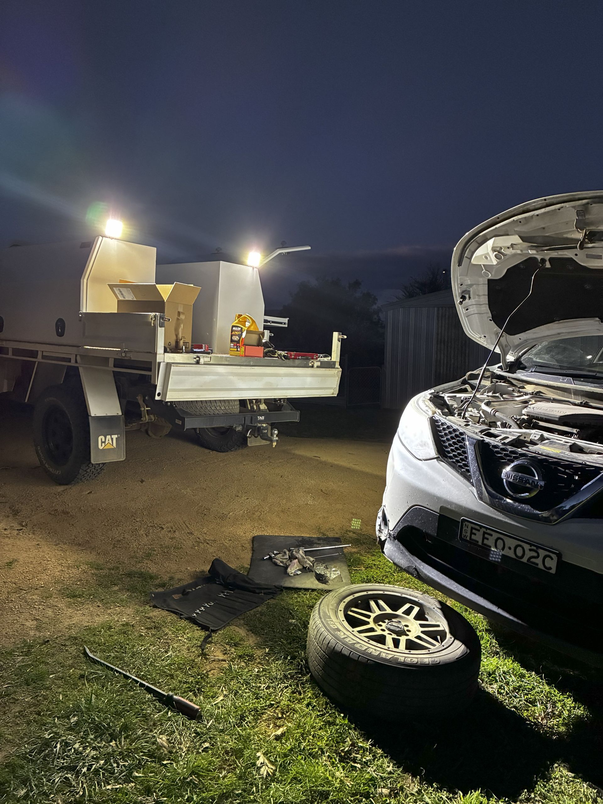 Car with open hood and flat tire being worked on, lit by trailer lights at night. — Bodes All Mechanical In Tathra, NSW