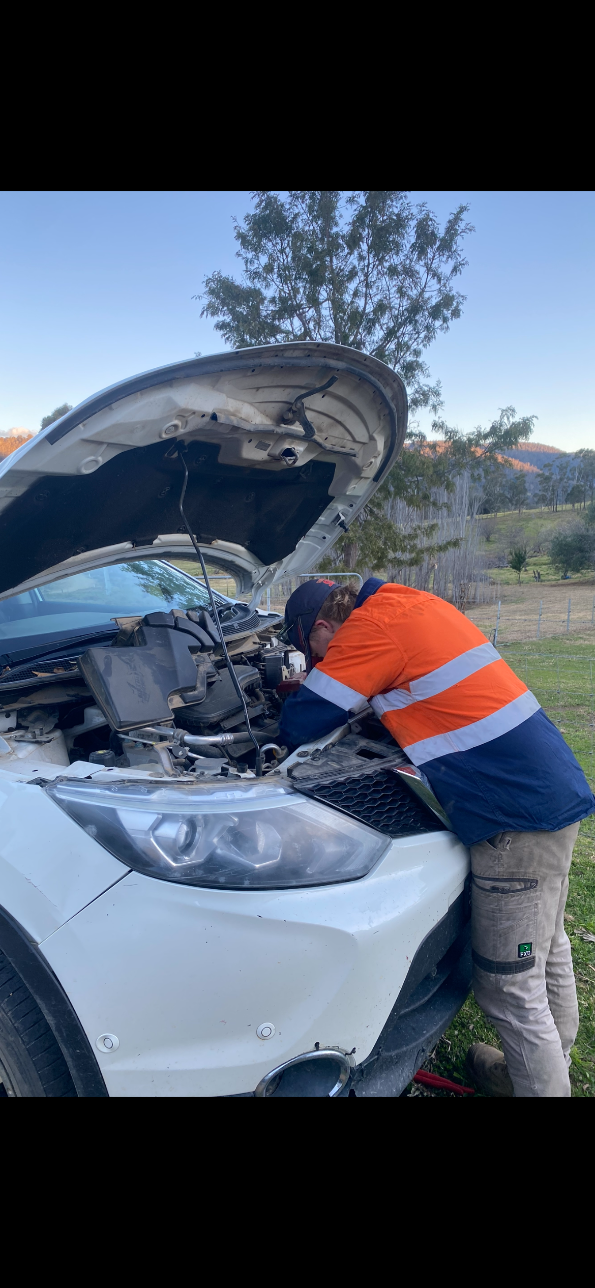 Person in orange work jacket working on the engine of a white car outdoors. — Bodes All Mechanical In Merimbula, NSW